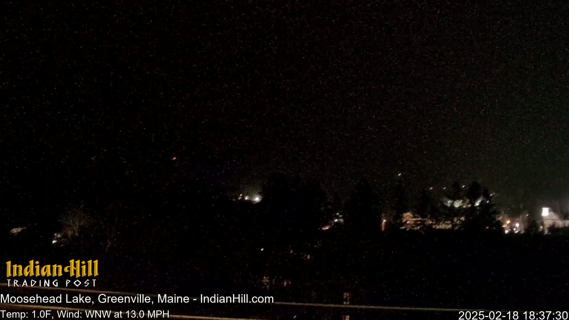 A nighttime view of a dark, snow-filled sky over a sparsely lit landscape with a wind of 13 mph.