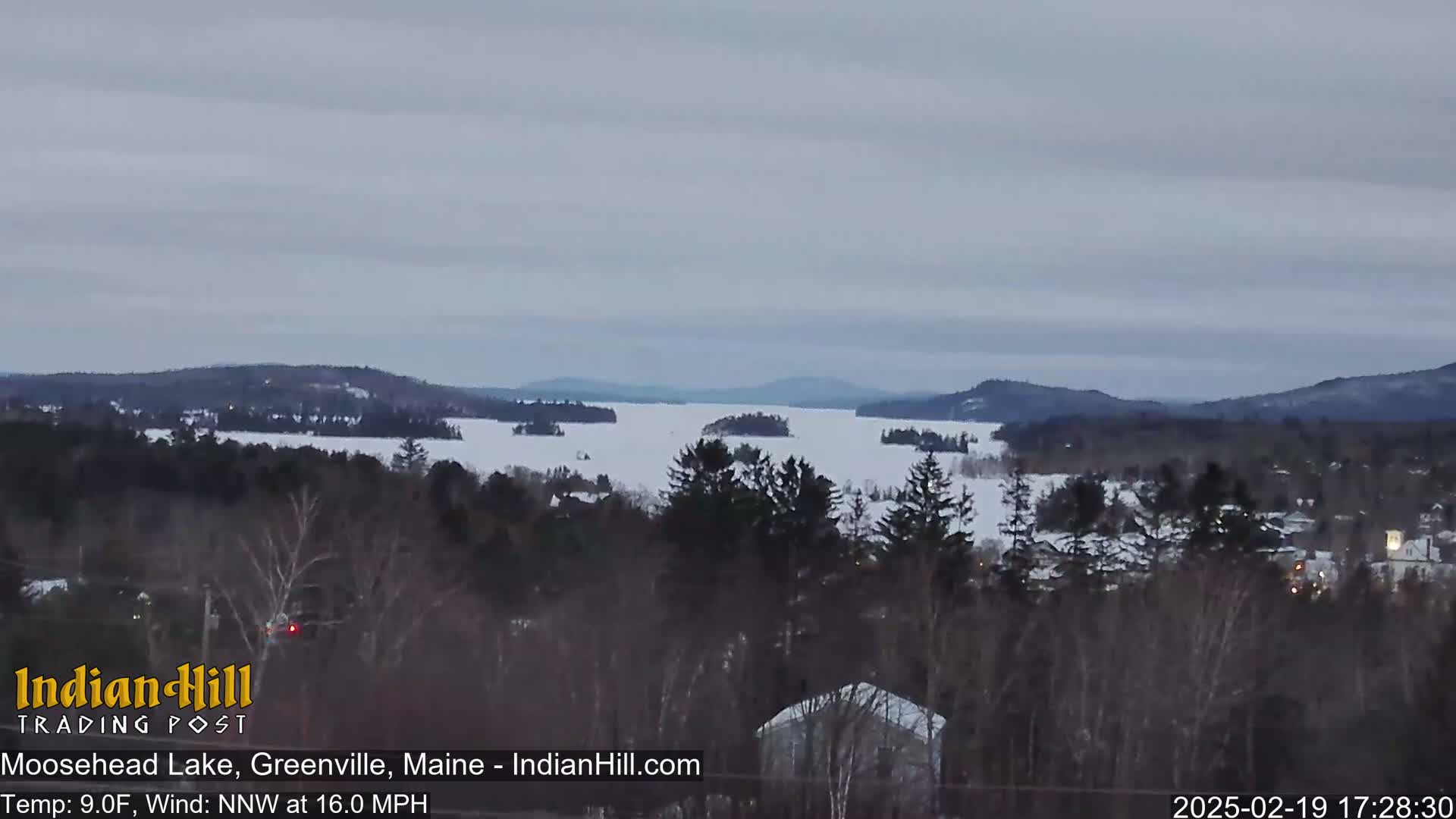 A mostly frozen lake stretches to snow-covered hills under an overcast sky.