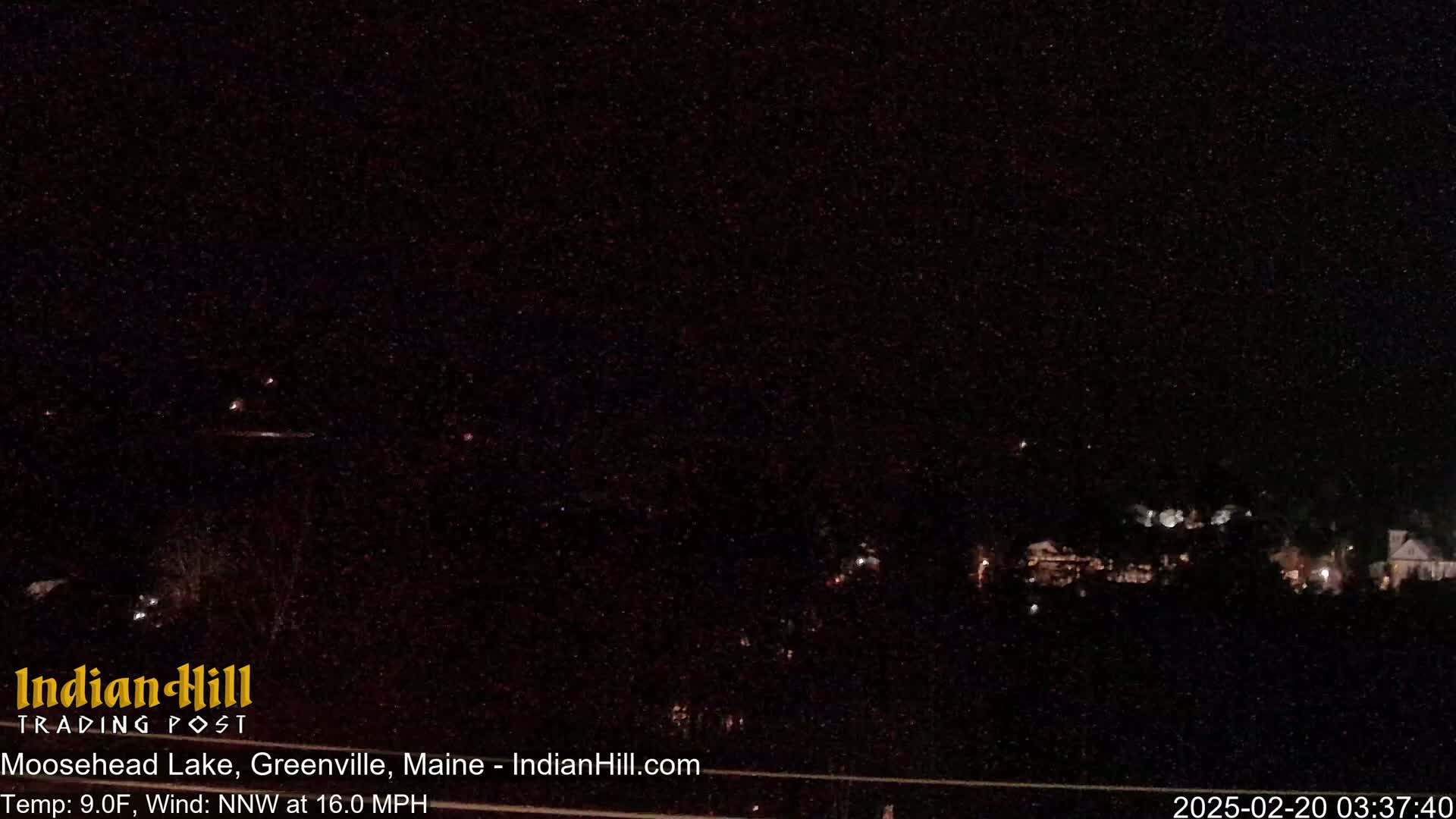 A nighttime, long shot of a dark, sparsely lit landscape with a few distant lights under a clear, starlit sky, with a 9°F temperature and a 16 mph north-northwest wind.