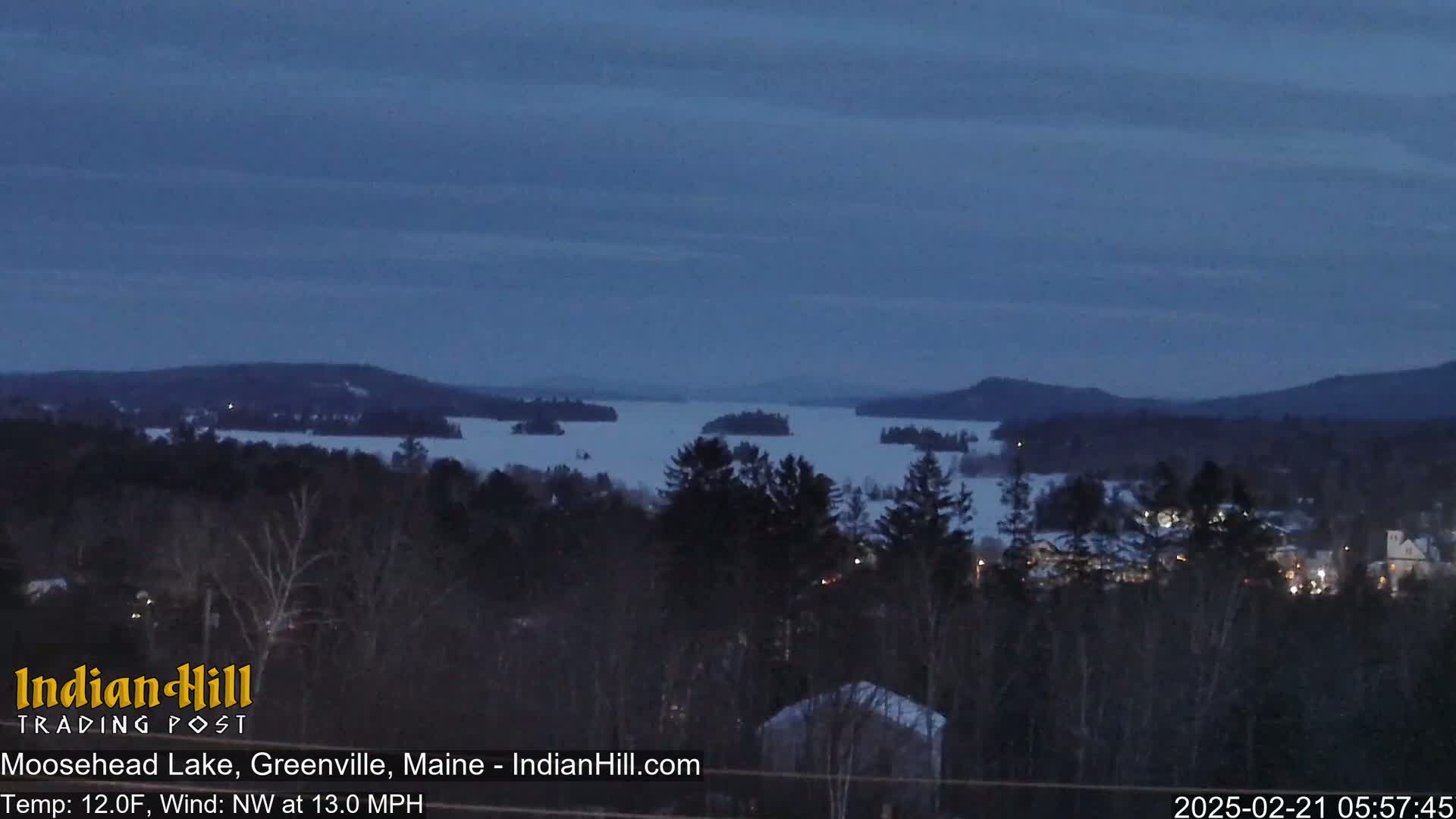 A twilight view of a snow-covered lake with several islands, surrounded by dark evergreen trees and distant hills under a mostly clear, dark blue sky.