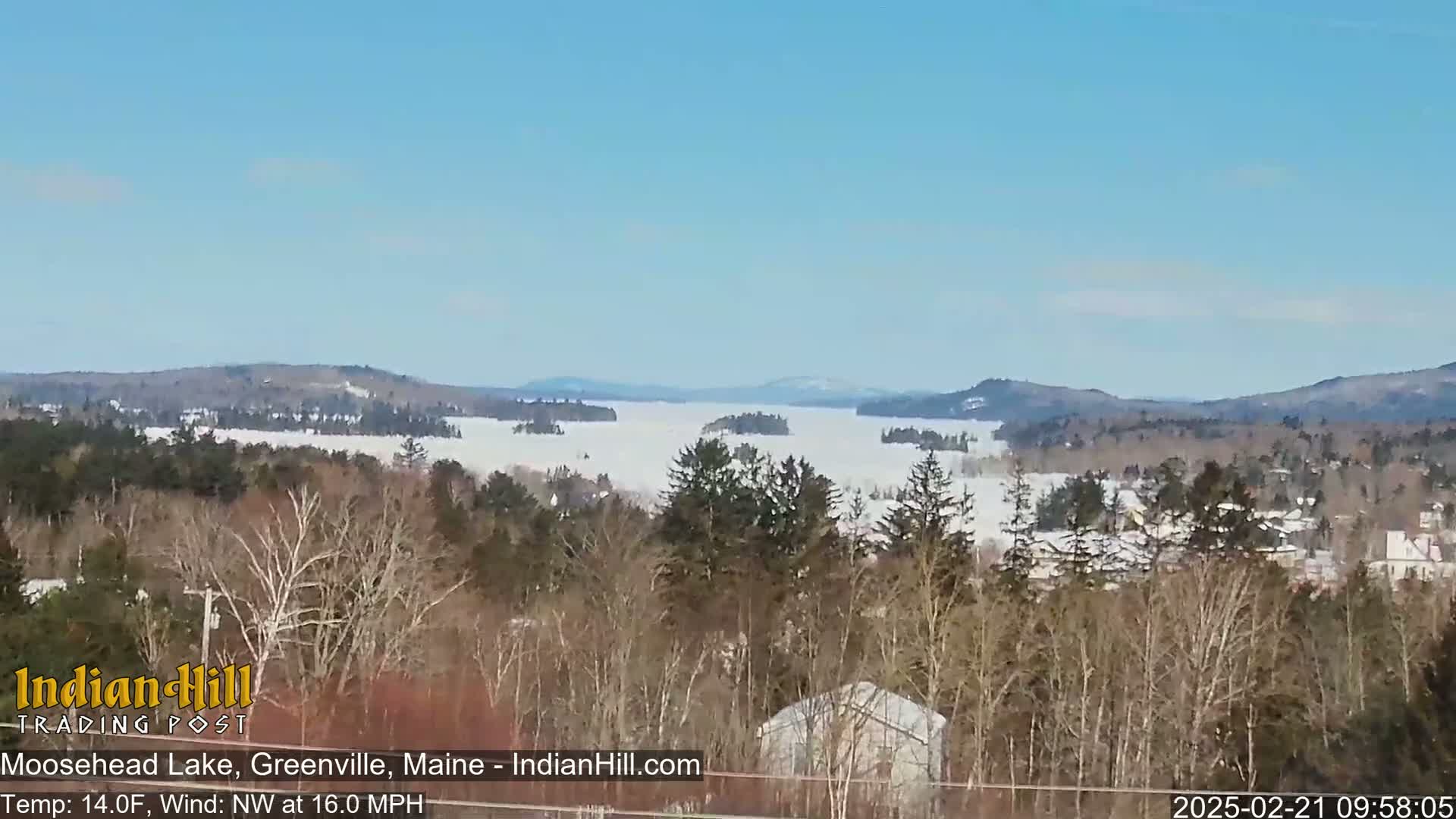 A snow-covered lake is visible in the distance, framed by bare winter trees and distant mountains under a clear blue sky, with a northwest wind at 16 mph.