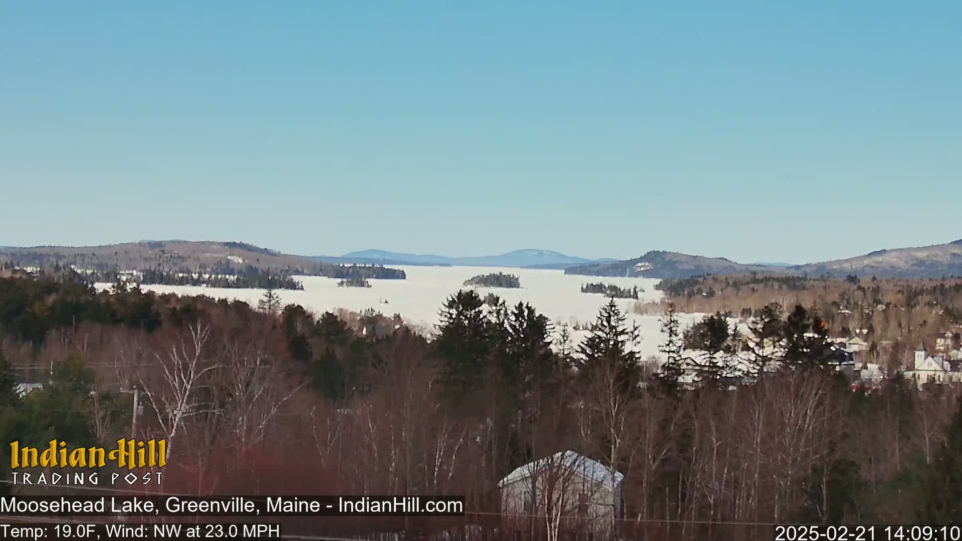 A snow-covered lake stretches to distant mountains under a clear blue sky, with a forest of leafless trees in the foreground.