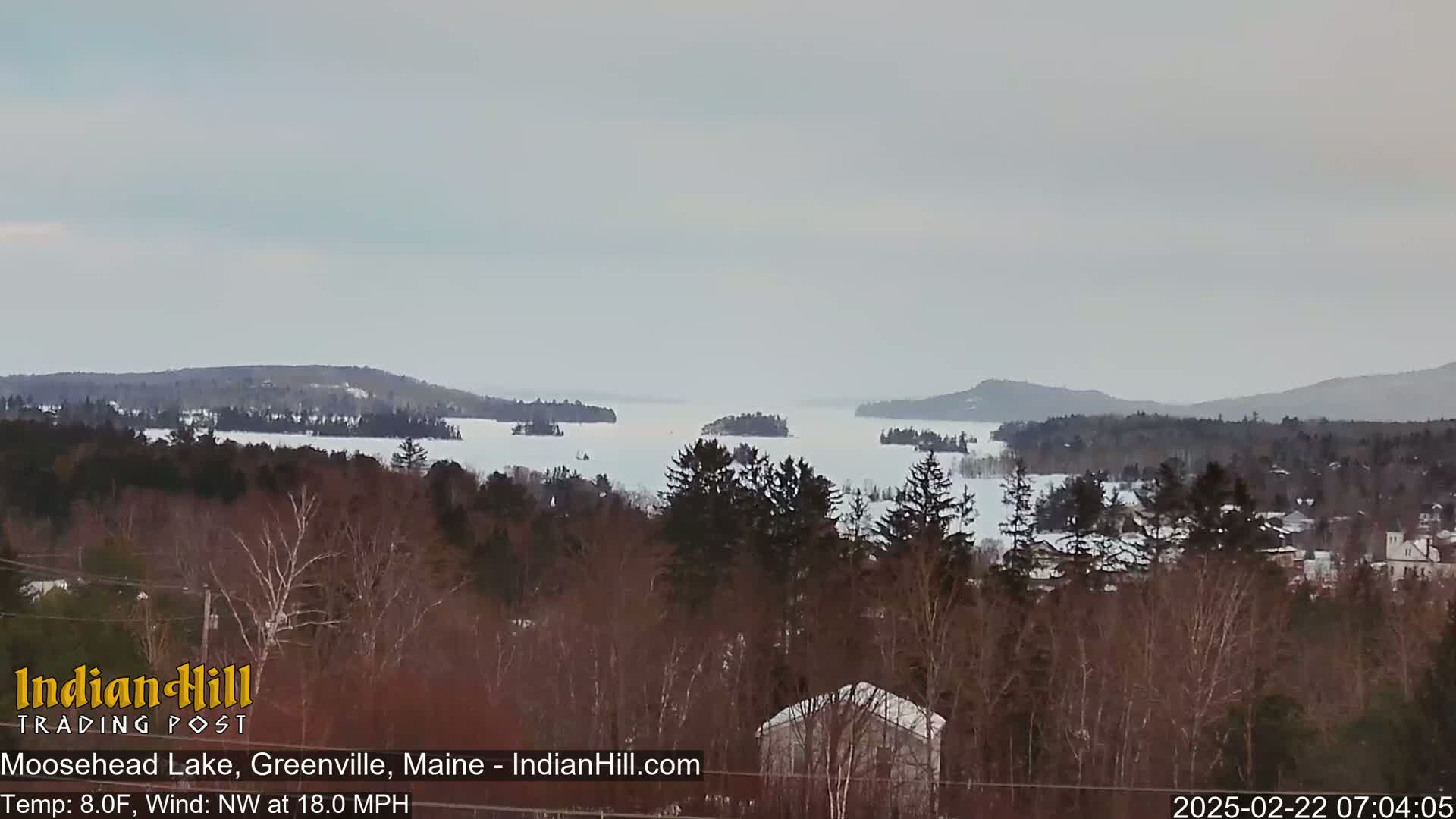 A partly frozen lake is visible from a snowy, wooded hillside on a cloudy, cold day with a northwest wind.