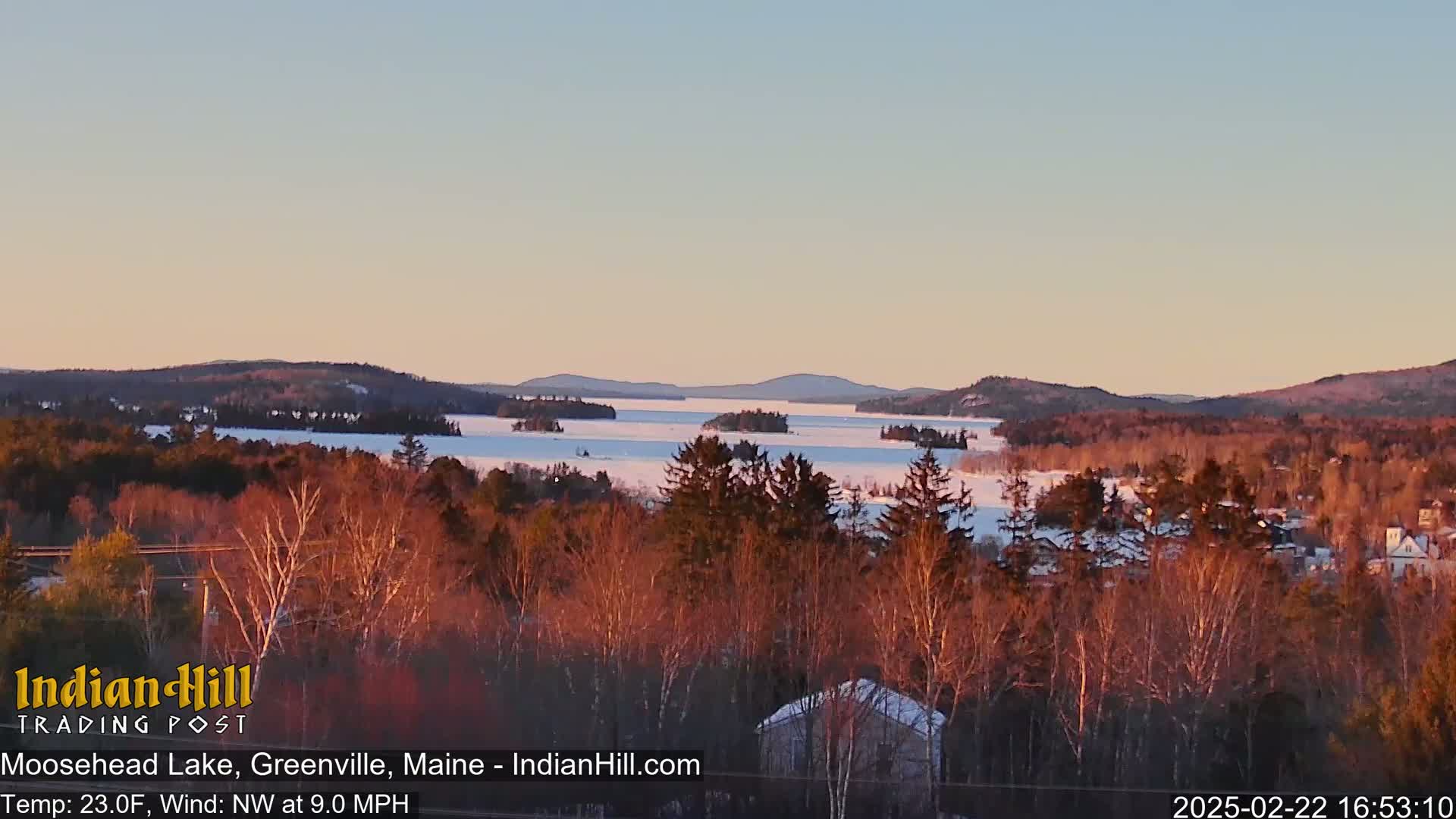A partially frozen lake is seen from a distance, surrounded by snow-covered trees and hills under a clear, soft pink and pale orange sunset.