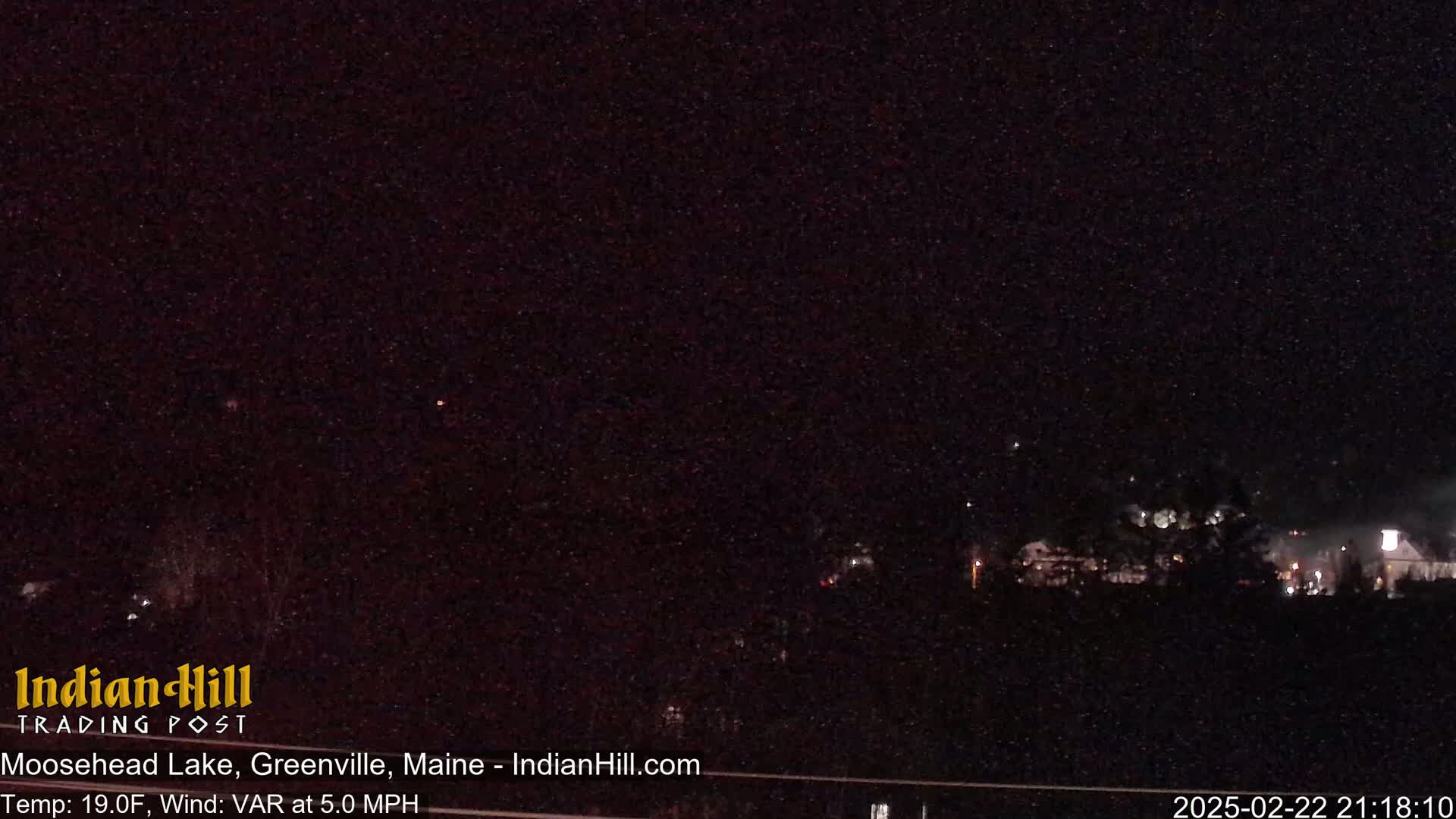 A nighttime long shot of a dark landscape with a few faint lights in the distance under a clear, starry sky.