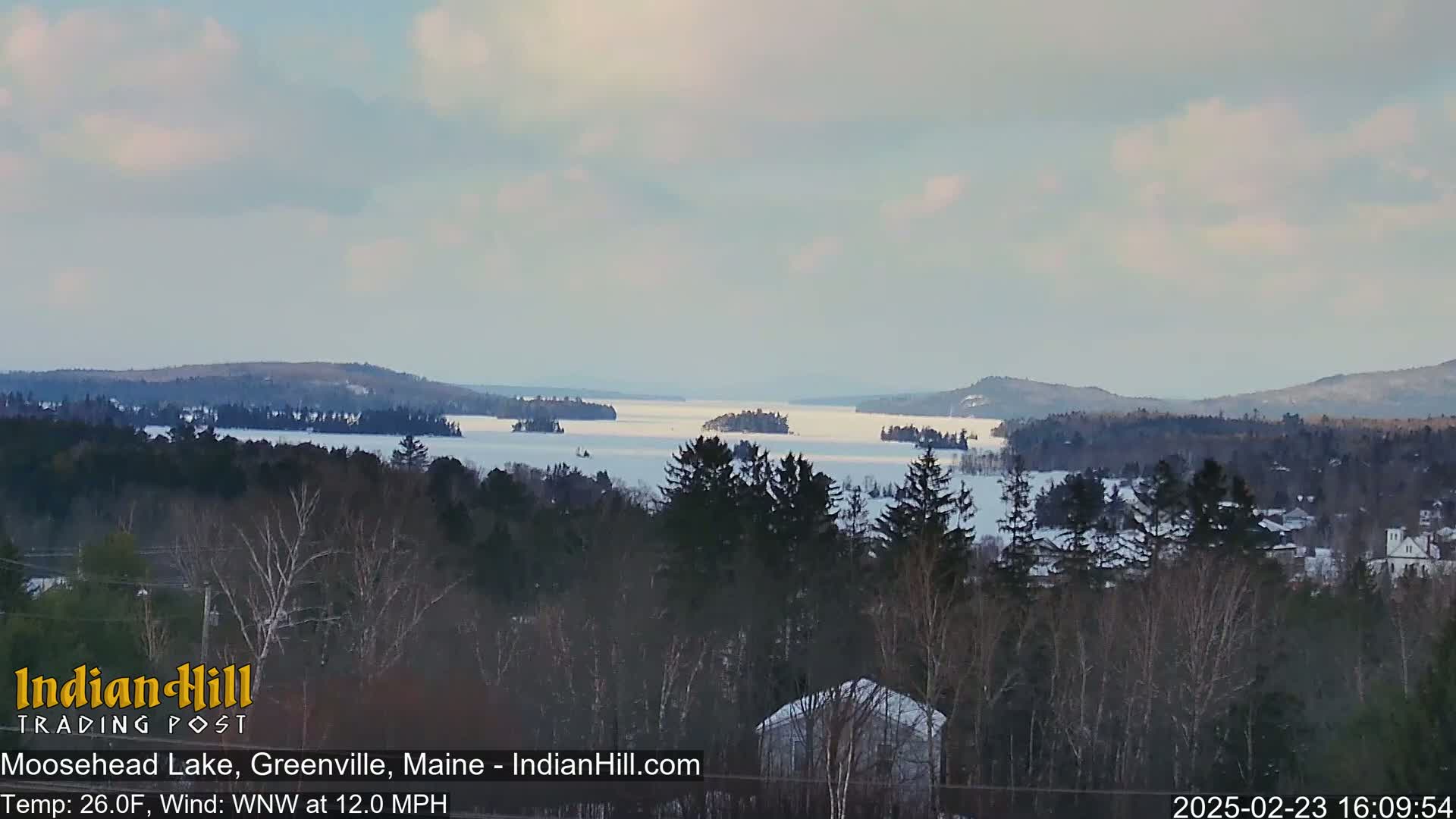 A snow-covered lake with several islands is seen from a snowy, wooded hillside under a mostly cloudy sky.