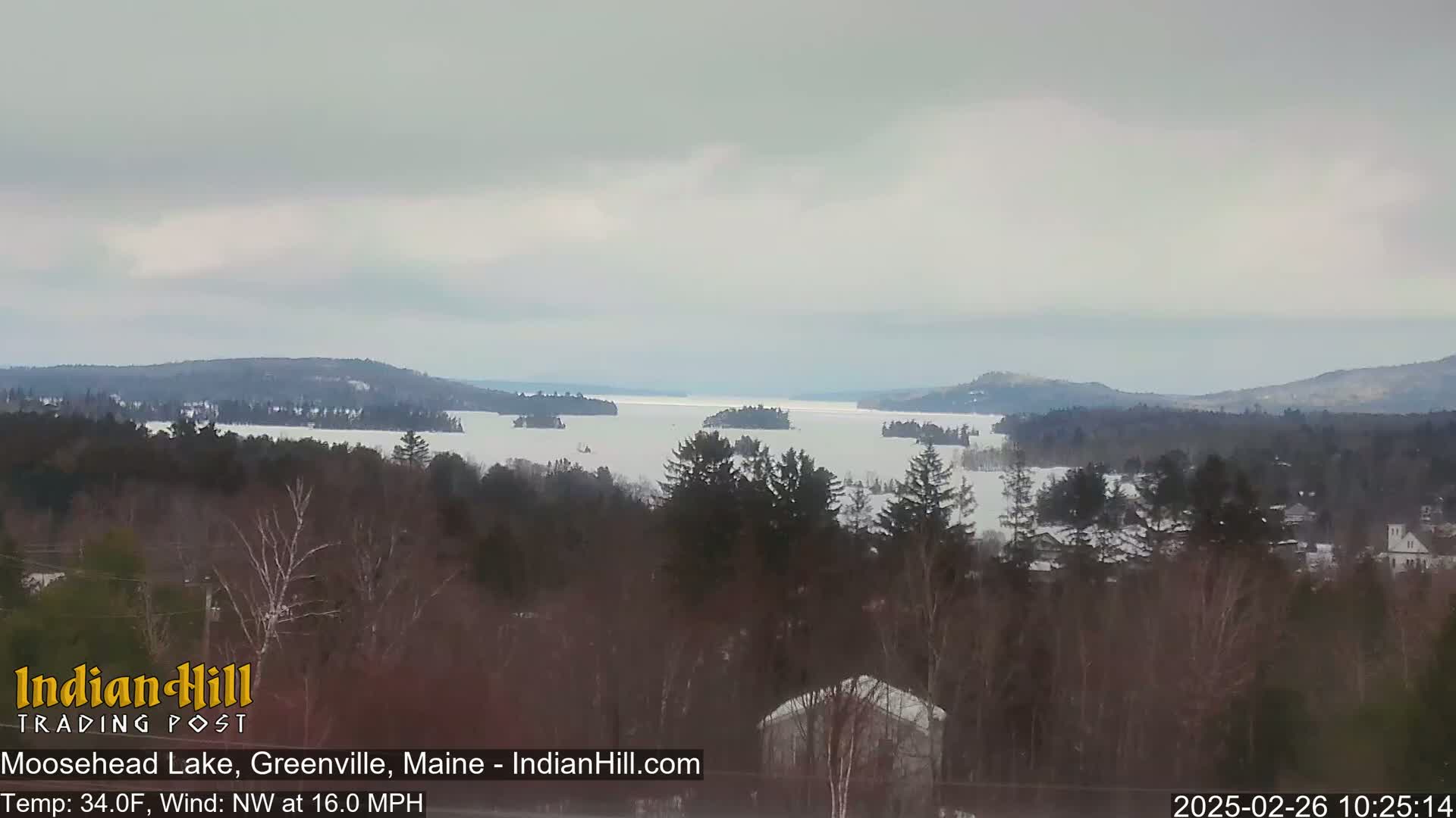A partially frozen lake is surrounded by snow-covered trees and hills under an overcast sky.