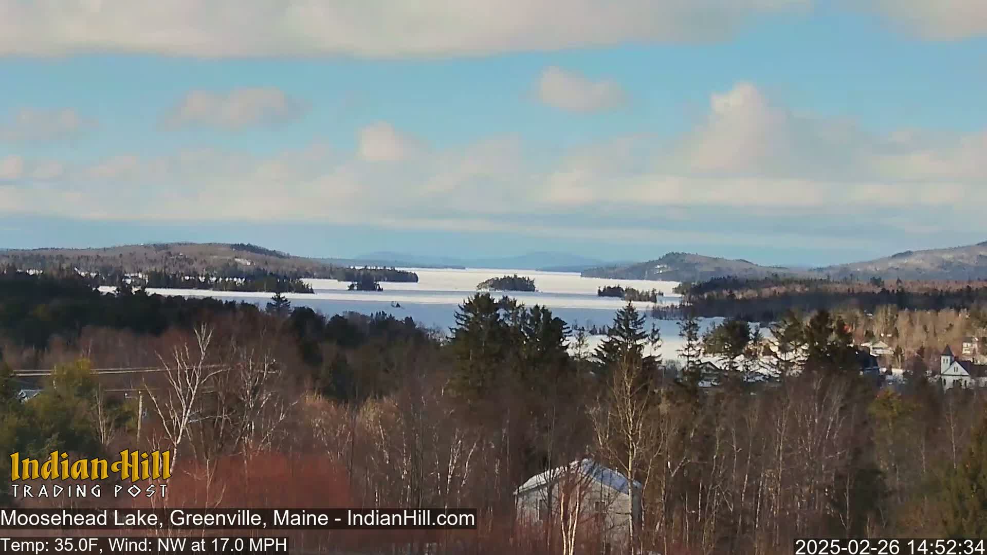 A partially frozen lake is visible from a snowy, wooded hilltop under a partly cloudy sky.