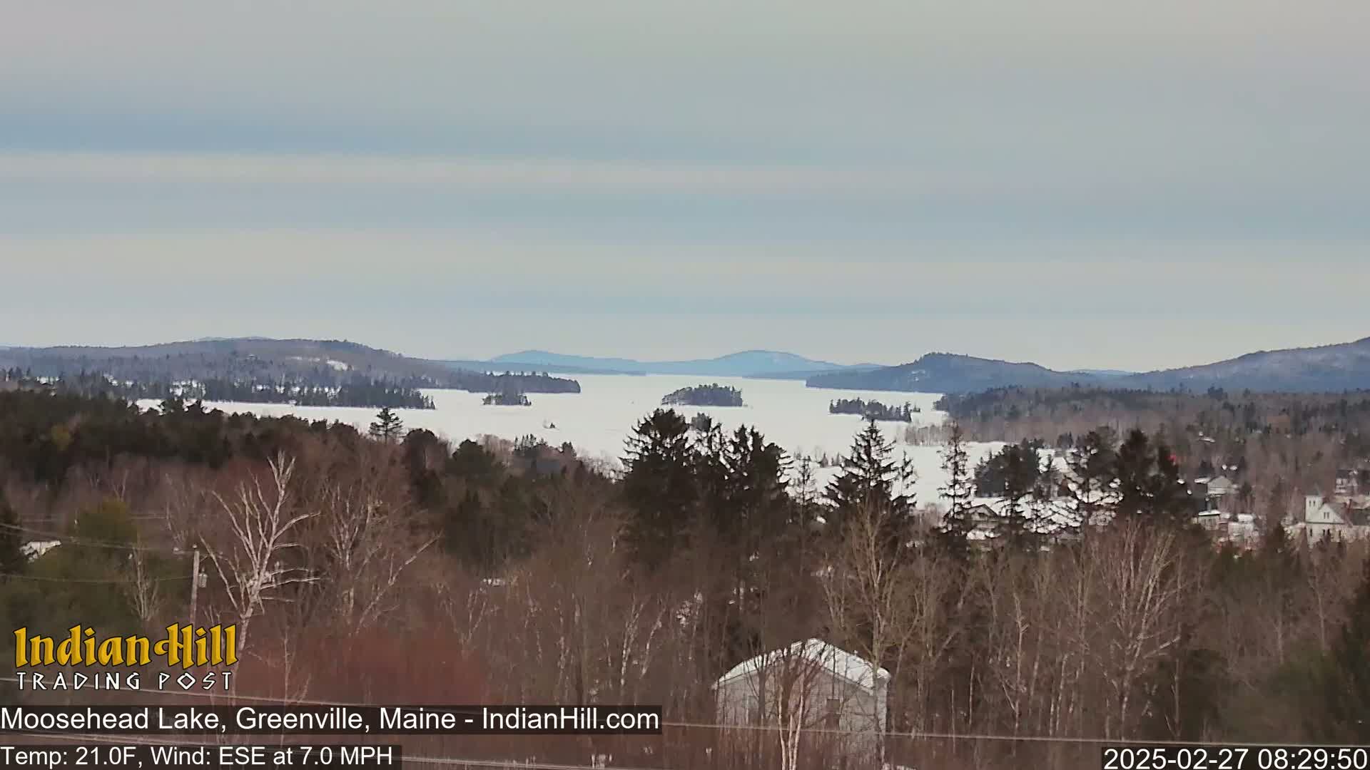 A snow-covered lake stretches into the distance, surrounded by a winter landscape of dark evergreens and bare deciduous trees under a mostly cloudy sky.