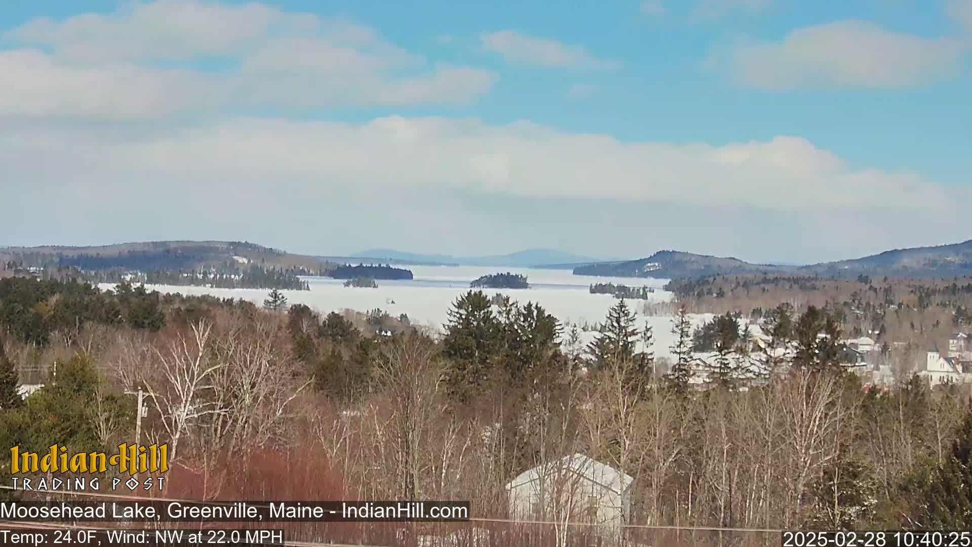 A snow-covered lake stretches to the horizon under a partly cloudy sky, viewed from a wooded hillside with a light northwest wind.