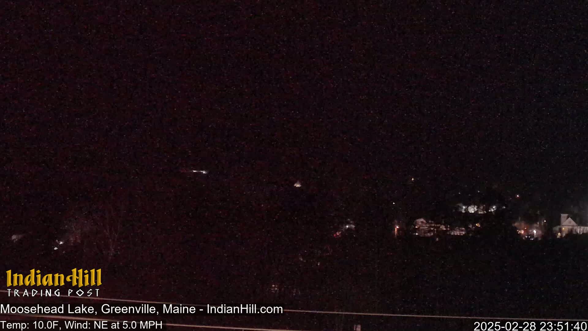 A nighttime long shot of a dark landscape with a few distant lights and a clear, starry sky.