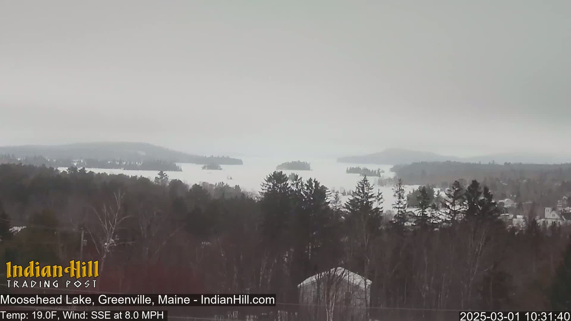 A snow-covered lake with several islands is visible through a hazy, overcast sky from a high vantage point overlooking a snow-covered forest.