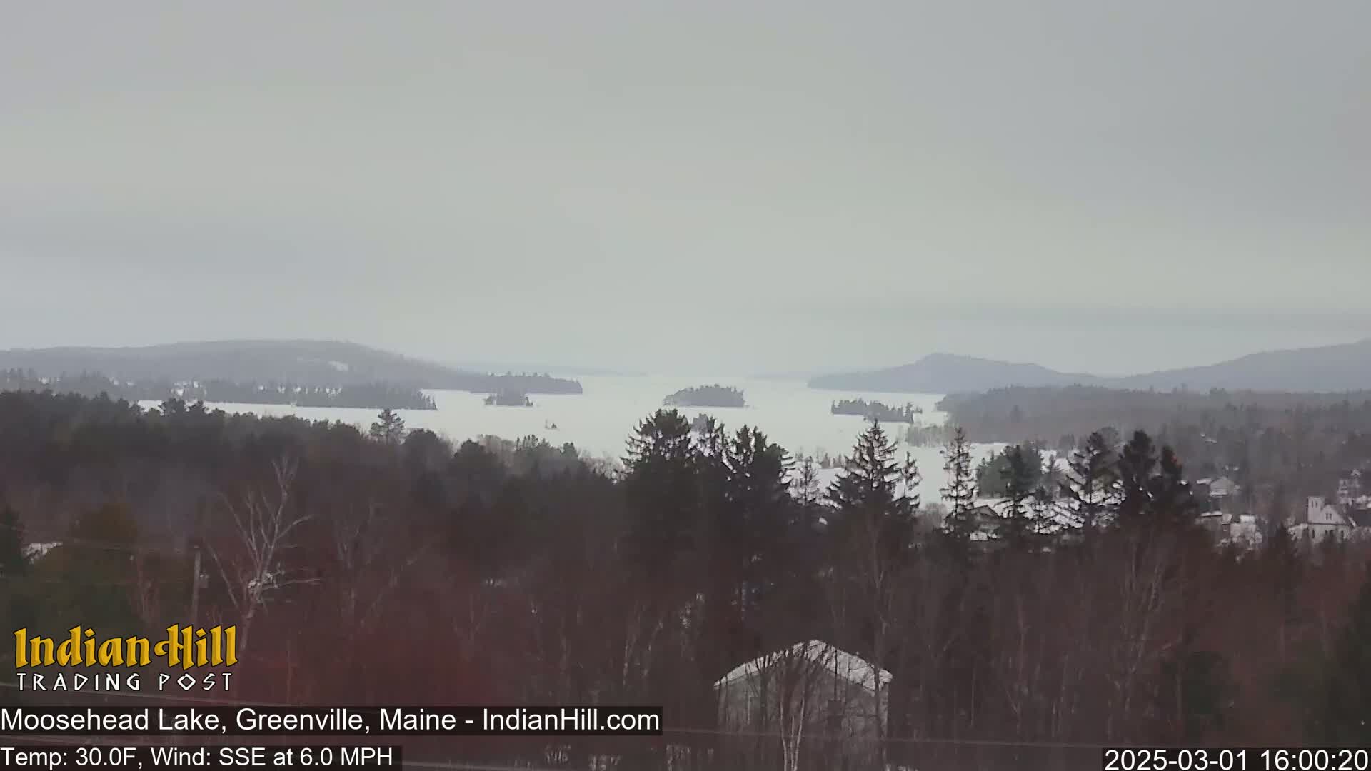 A mostly overcast, snowy landscape shows a large lake with several islands, surrounded by dark evergreen trees and distant hills.