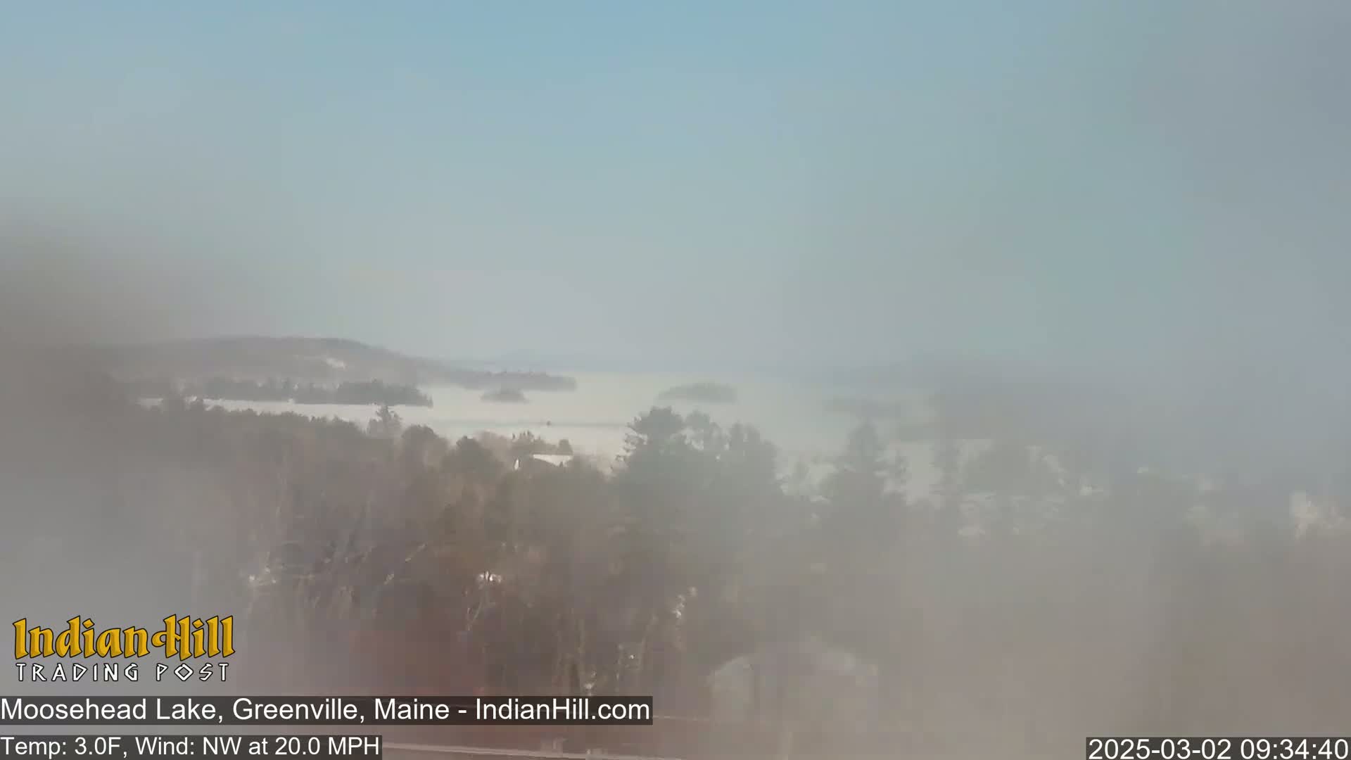 A foggy, snowy landscape shows a lake partially obscured by fog, with trees and a hint of land in the background, under a mostly clear sky.