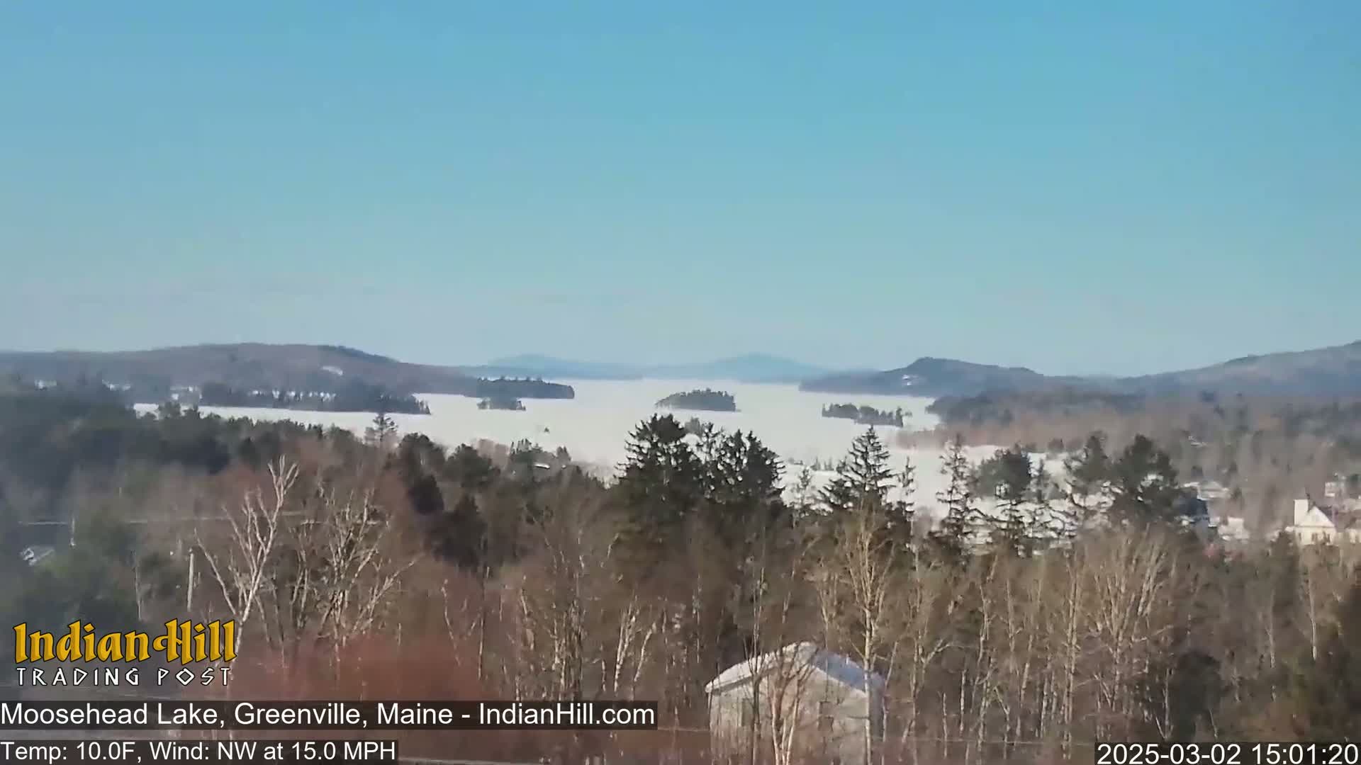 A partially frozen lake is visible from a snowy, wooded hillside under a clear blue sky, with a northwest wind blowing at 15 mph.