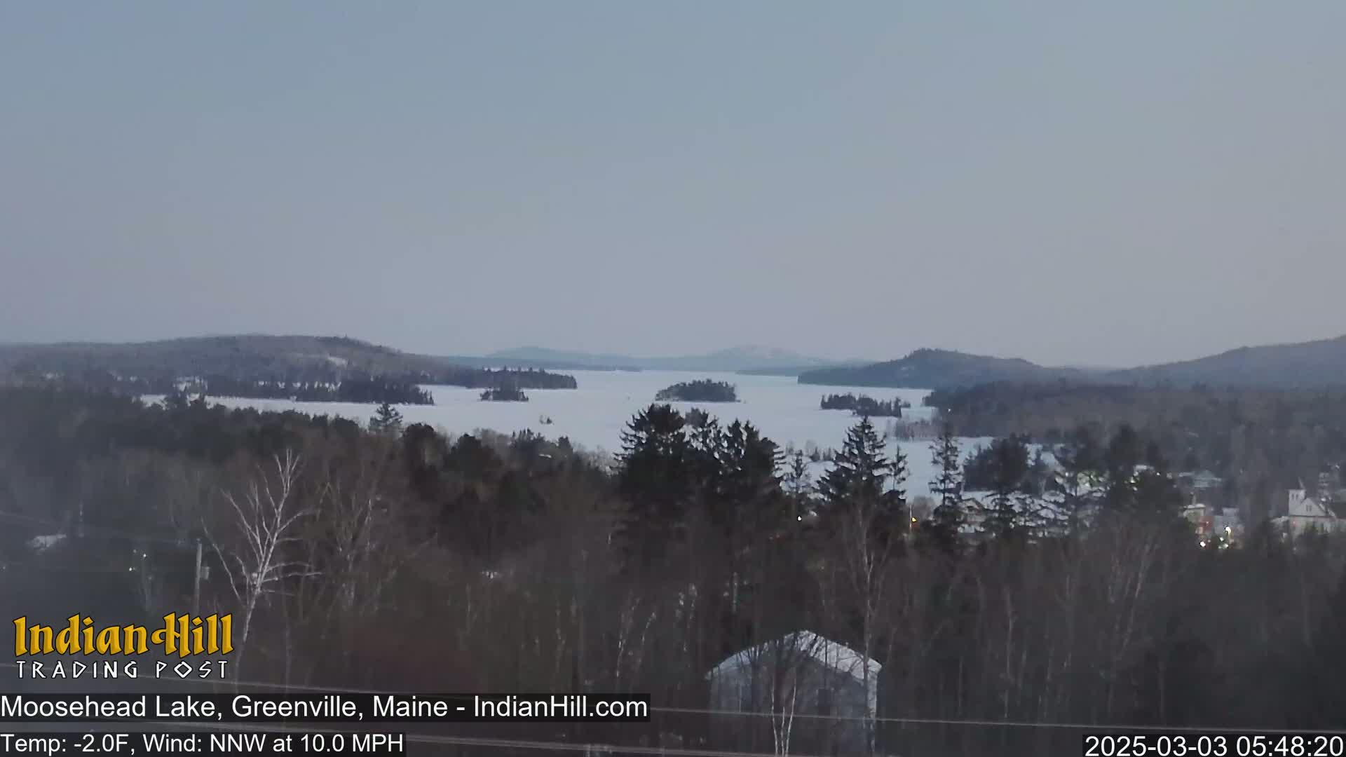 A snow-covered lake with numerous islands is visible from a high vantage point, with a chilly, windy, and overcast sky.