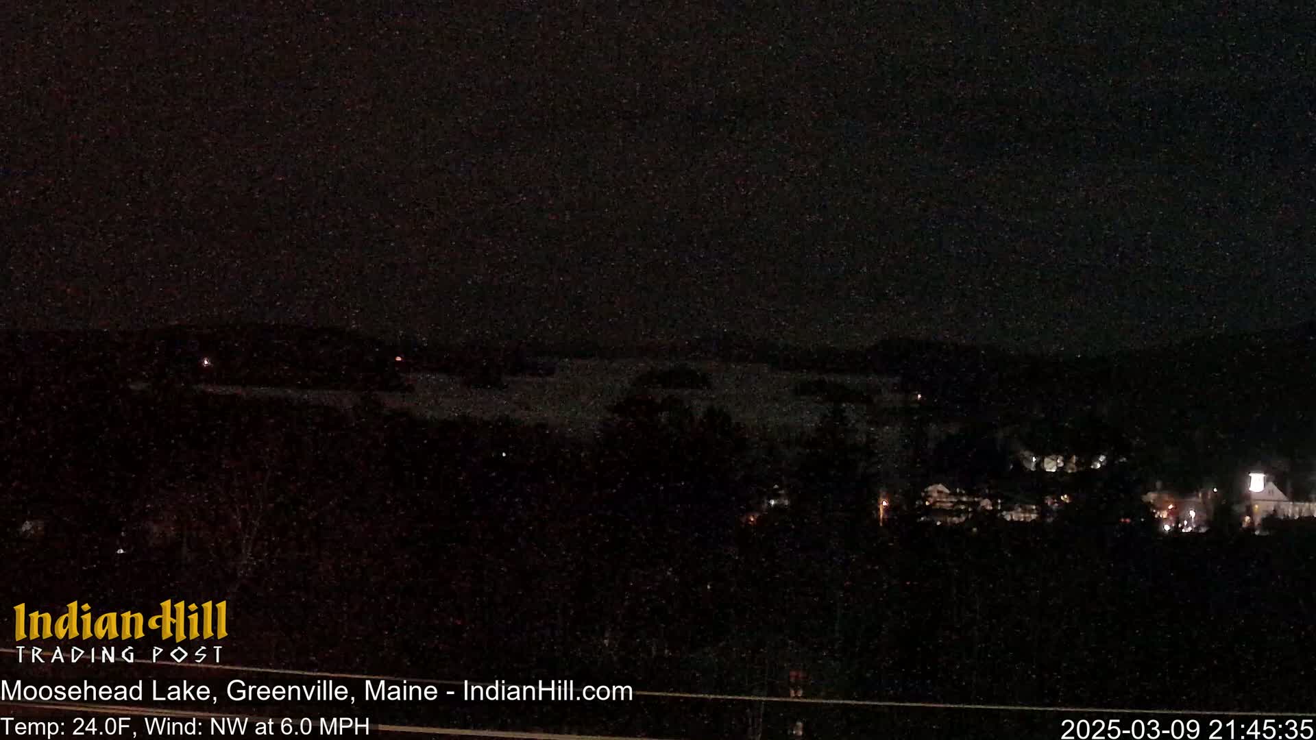 A dark nighttime view of a lake with a town visible in the distance under a clear sky.