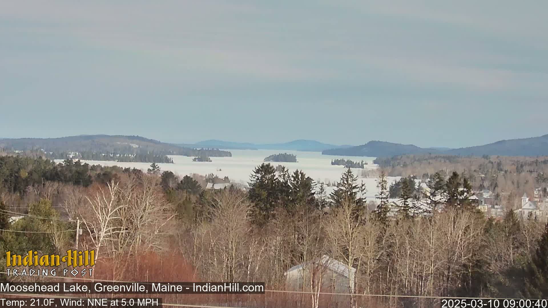 A snow-covered lake stretches to distant, tree-covered hills under a mostly clear sky.