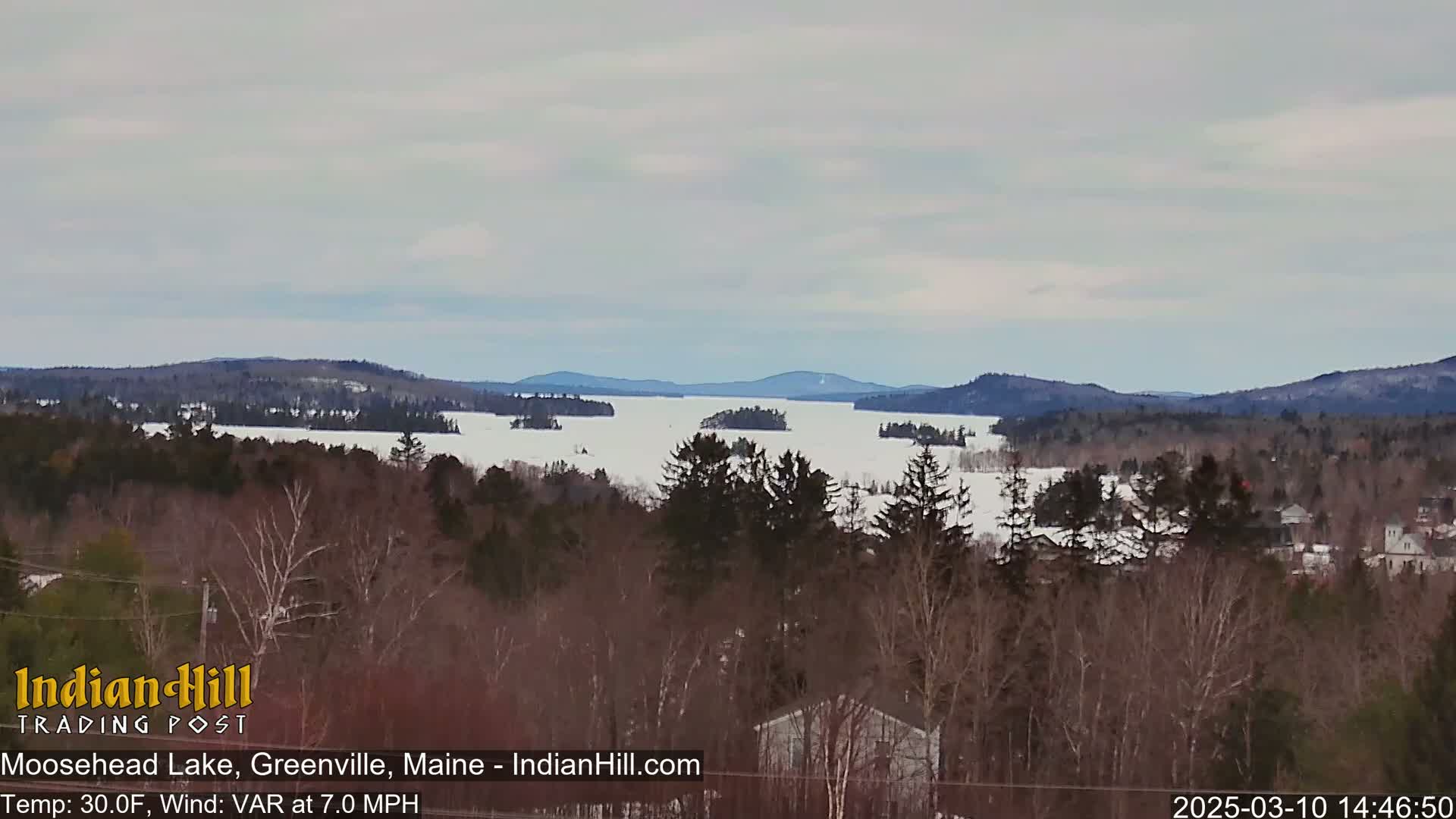 A snow-covered lake with several small islands is seen from a distance, surrounded by dark-colored, leafless trees under an overcast sky.