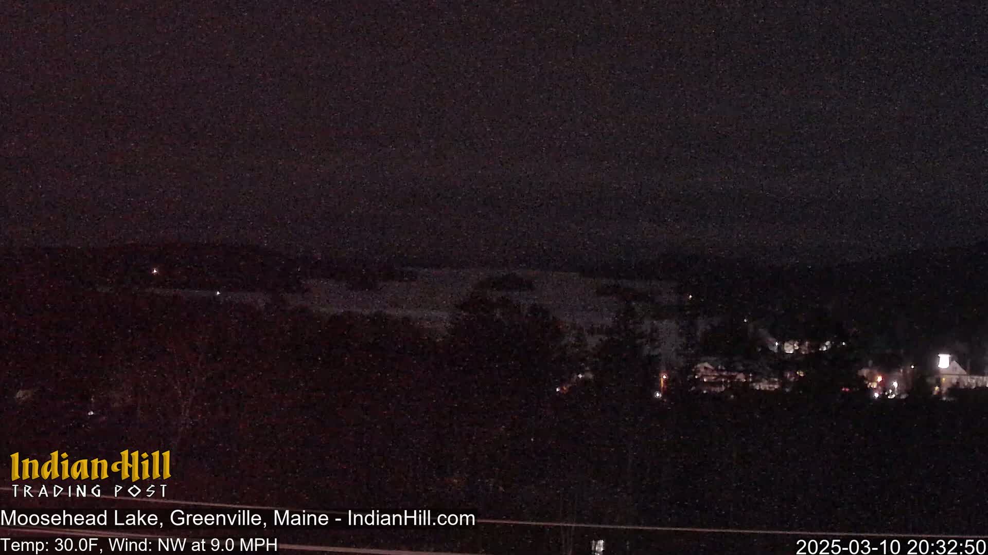 A dark, nighttime view of a lake and distant buildings under a clear sky with a northwest wind at 9.0 mph and a temperature of 30 degrees Fahrenheit.