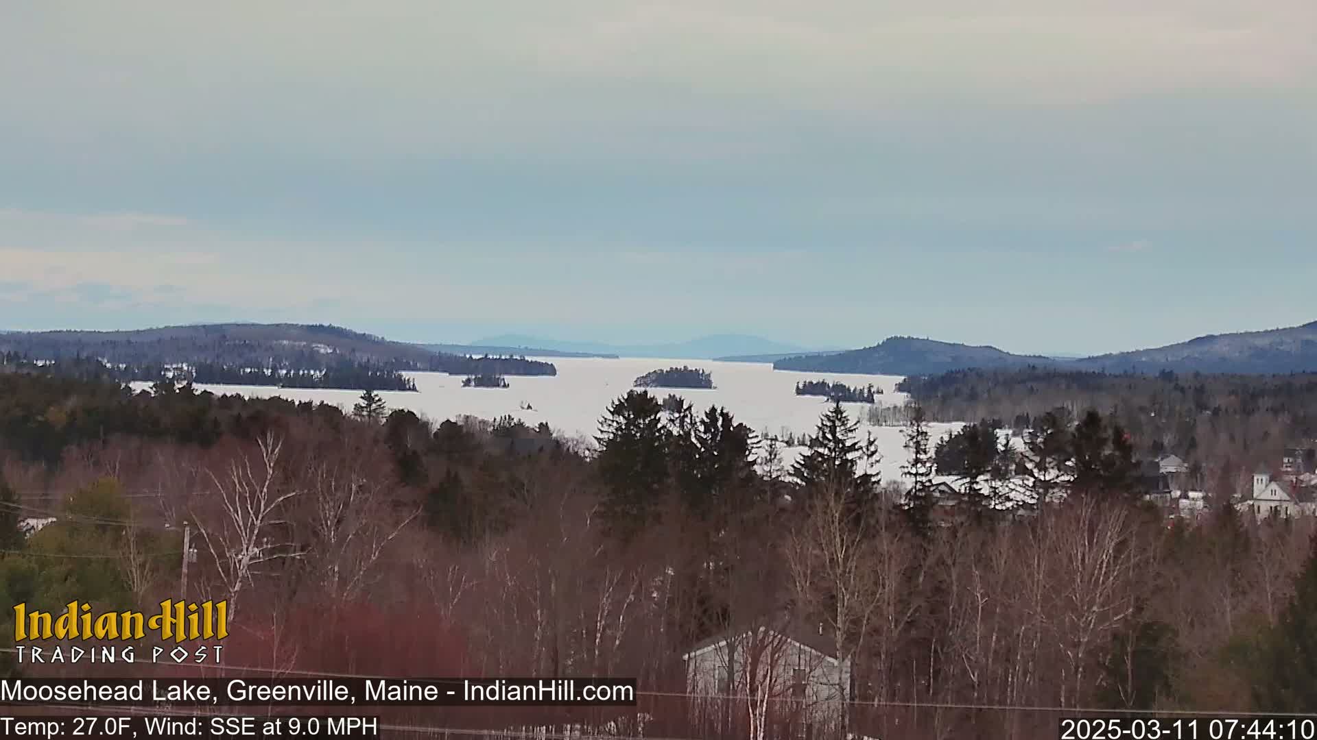 A snow-covered lake, surrounded by dark, leafless trees and distant mountains, is viewed from a high vantage point on a partly cloudy day with a south-southeast wind.