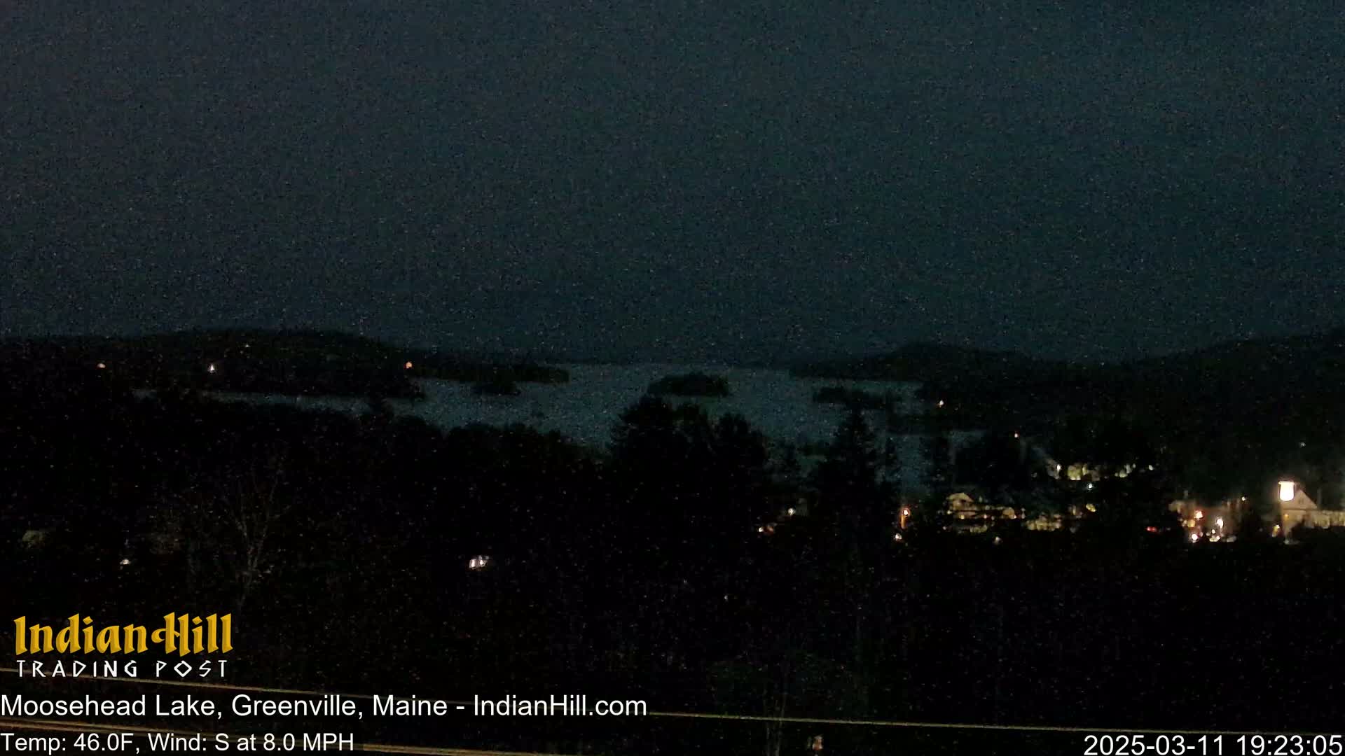 A dark, nighttime view of a lake and a small town under a slightly snowy sky.