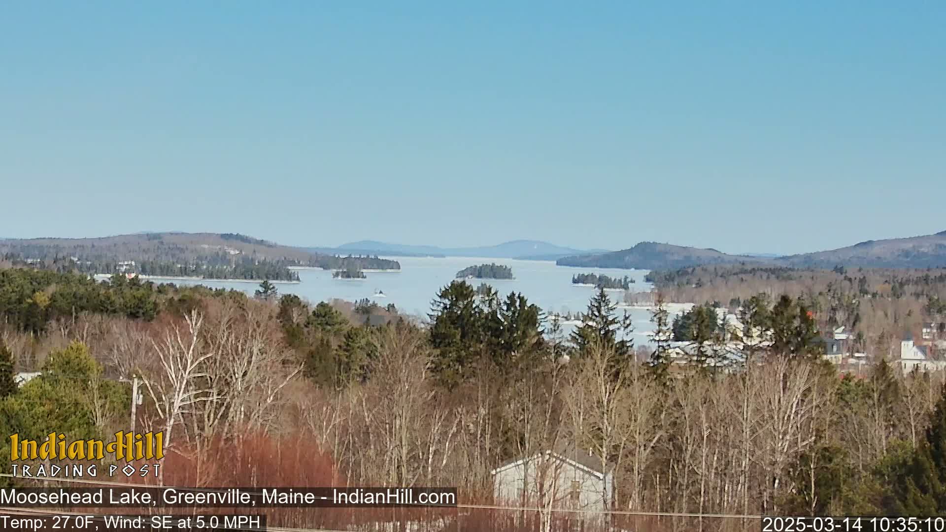 A partially frozen lake is visible from a wooded hilltop on a clear, sunny day.