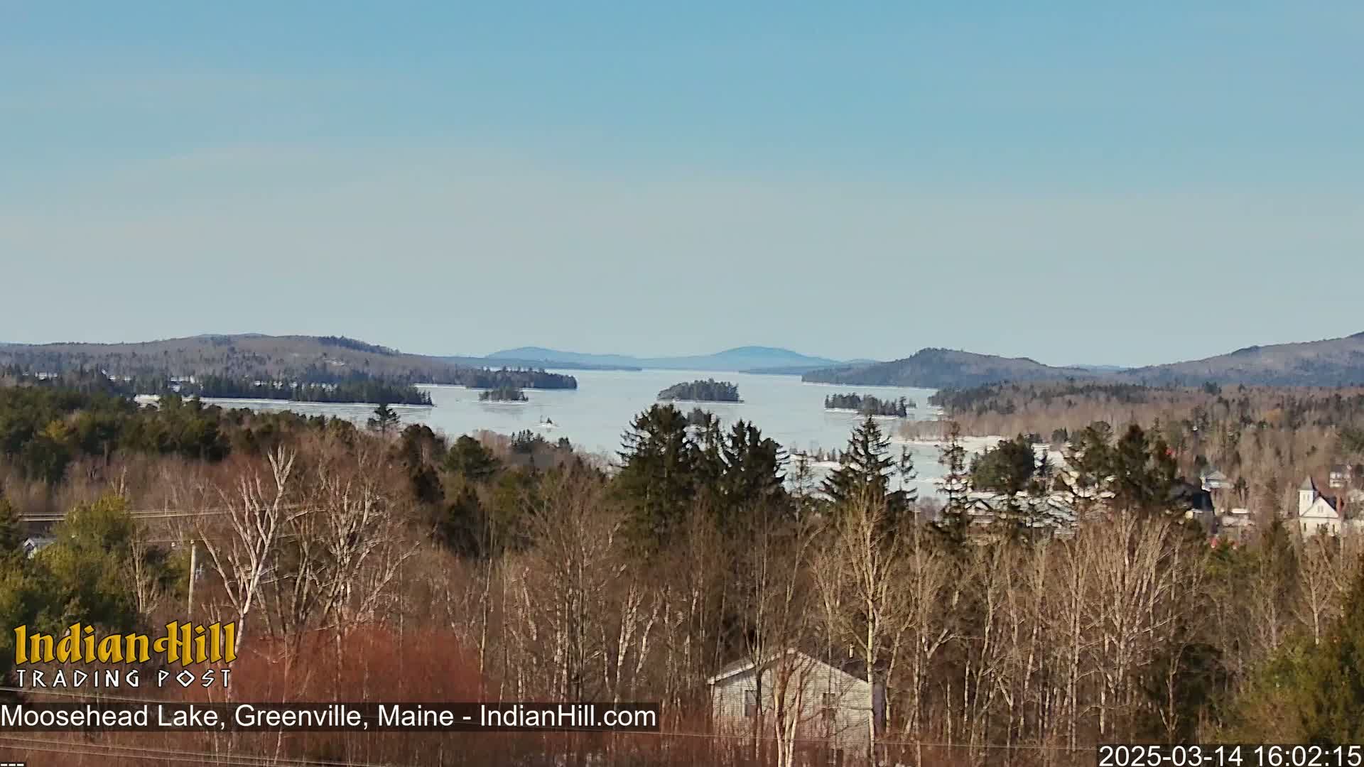 A partly frozen lake is visible from a wooded hillside under a clear blue sky.
