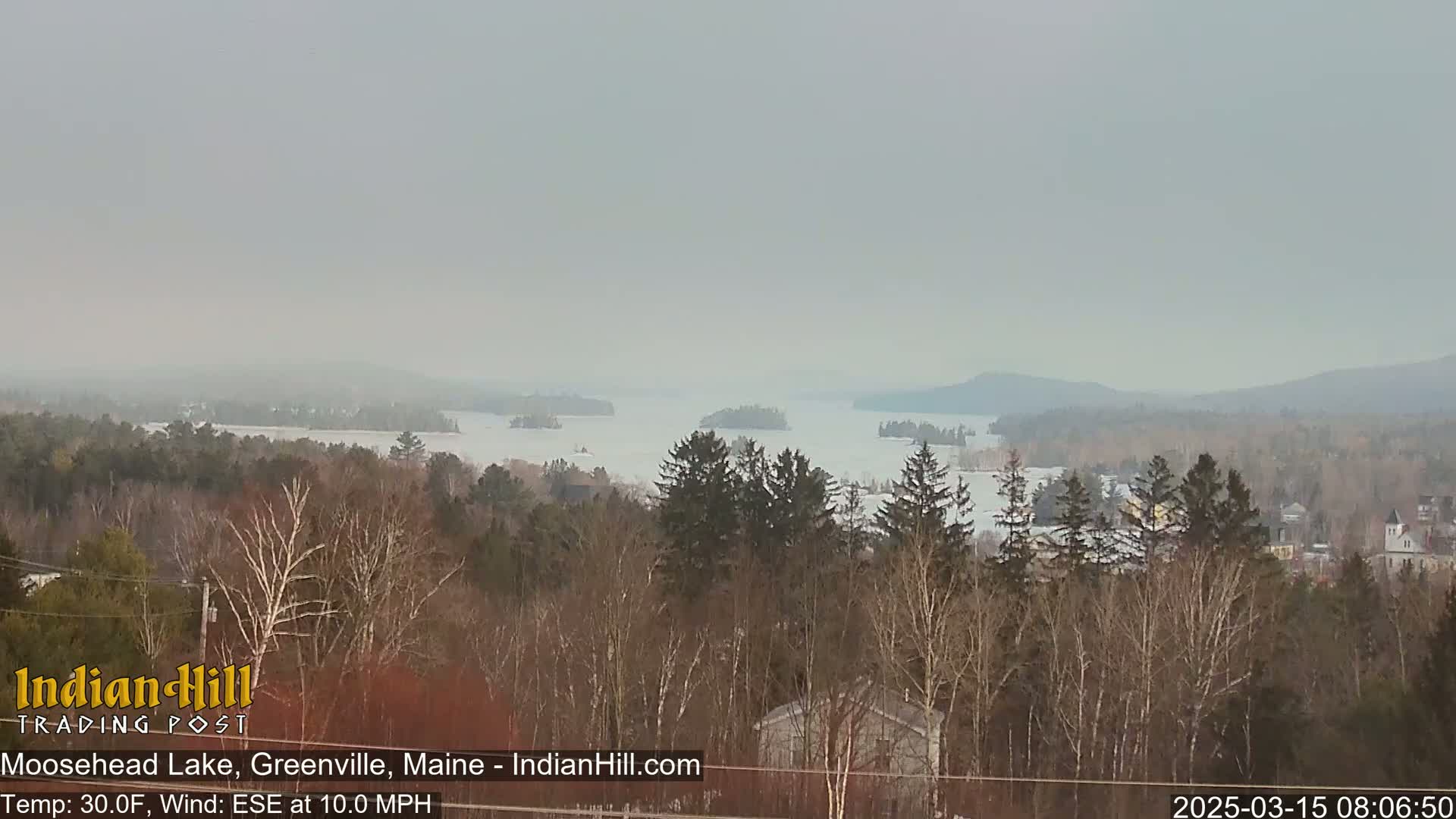 A hazy, cold view of a partially frozen lake surrounded by snow-covered trees and distant hills under an overcast sky.