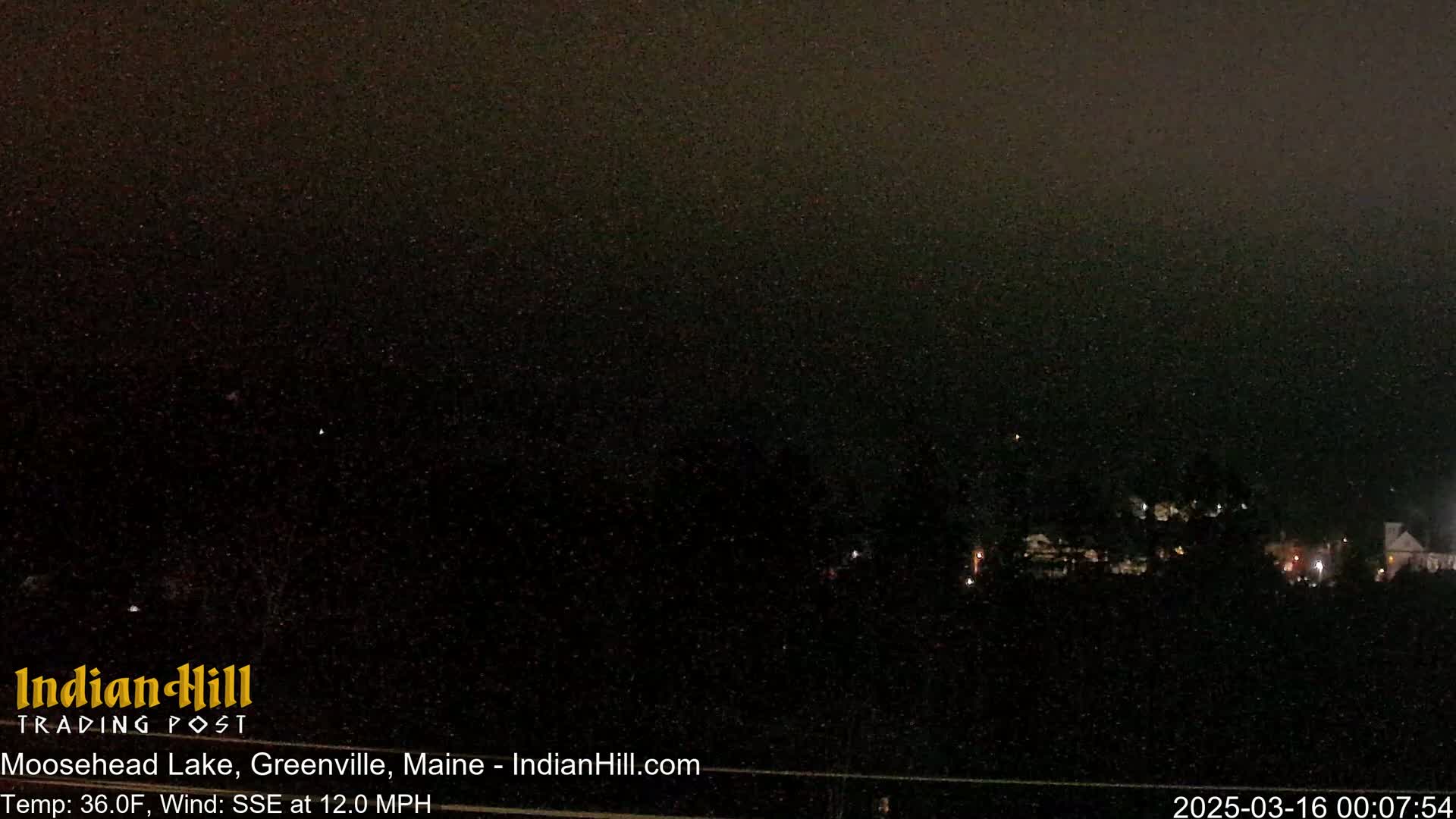 A nighttime long shot of a dark landscape with a few distant lights under a dark, possibly snowy sky.