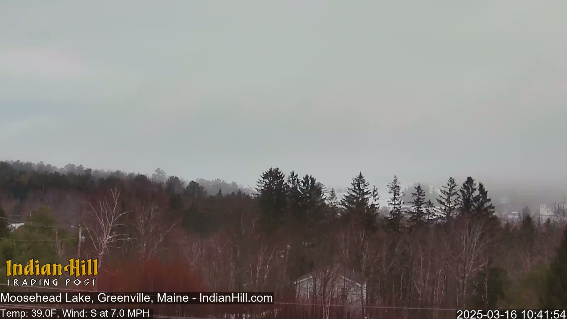 A mostly cloudy, somewhat hazy day with a view of a snow-dusted forest and a lake in the distance.