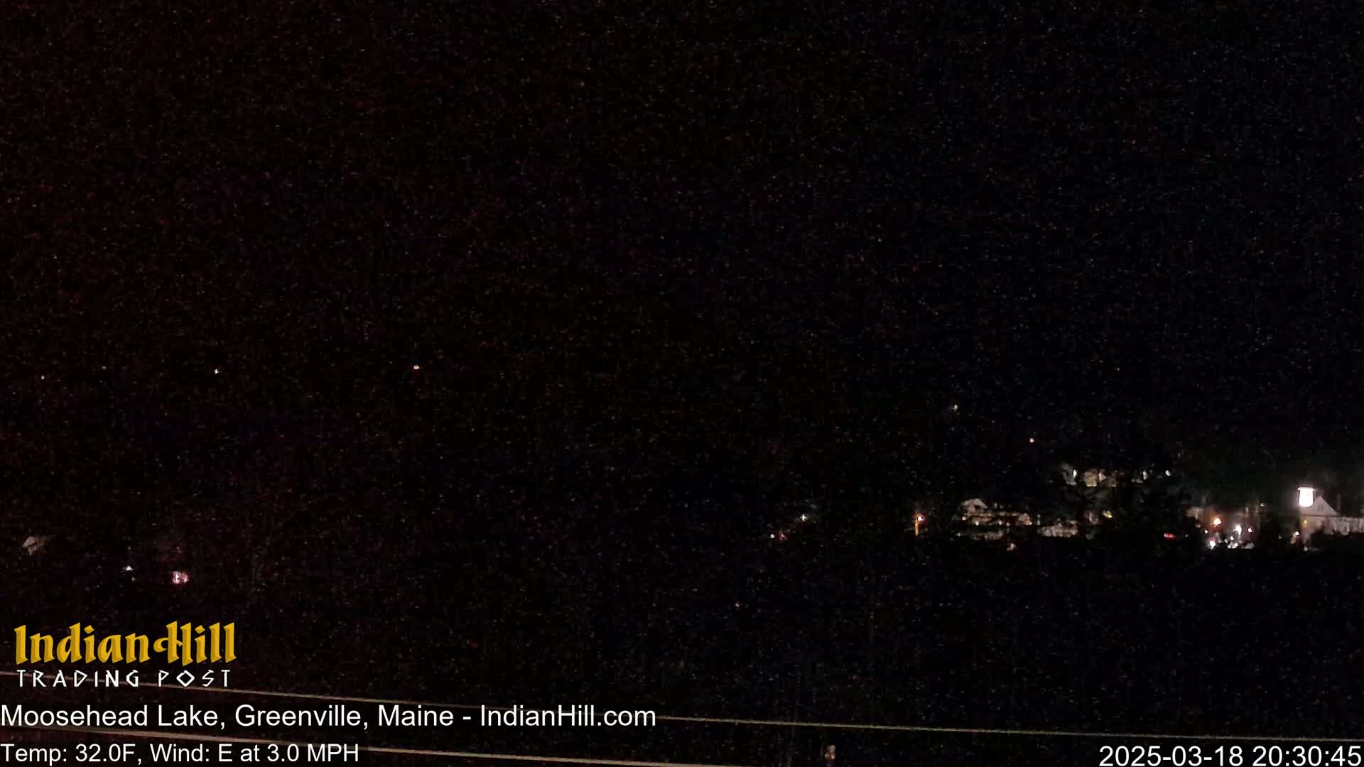 A nighttime, low-light view of a sparsely lit landscape with a few distant buildings under a dark, starry sky.