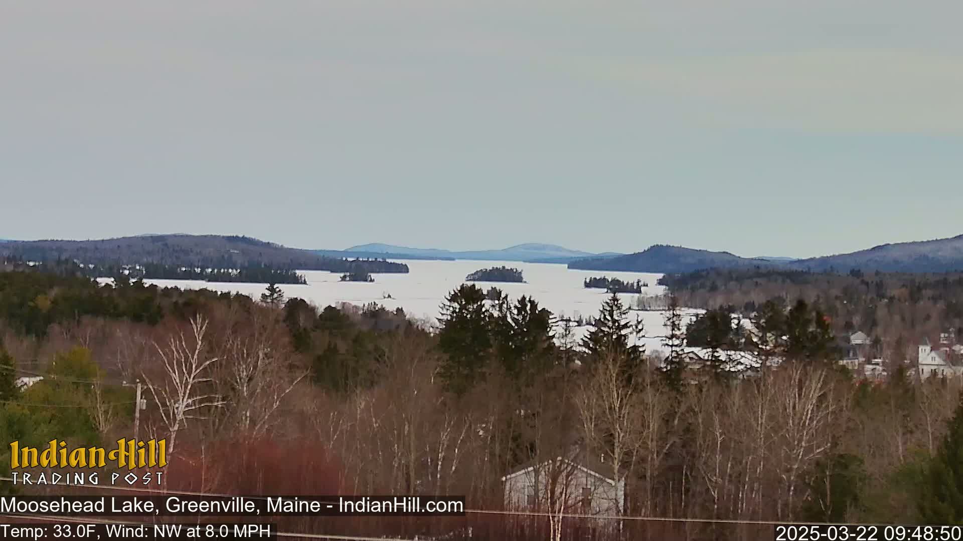 A partially frozen lake is seen from a high vantage point, surrounded by snow-covered trees and distant mountains under an overcast sky.