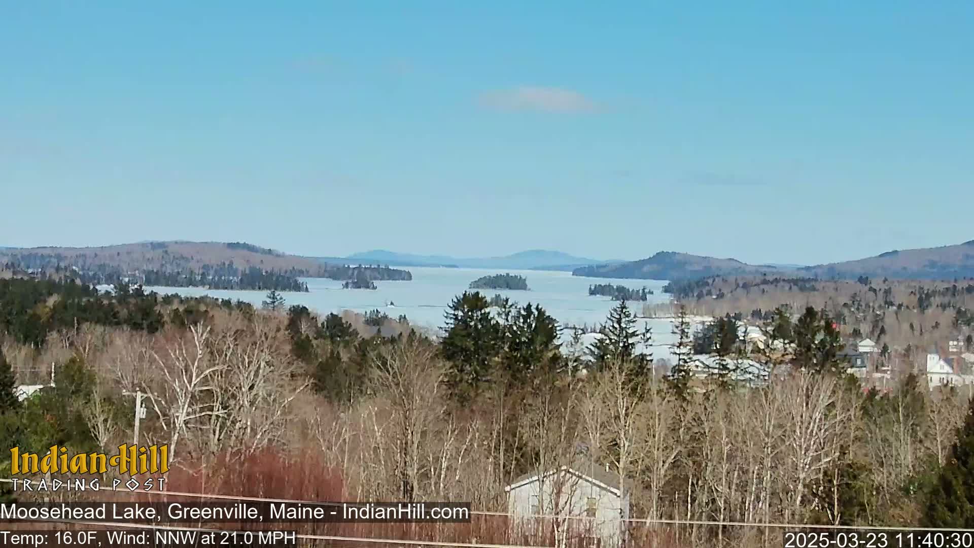 A partly frozen lake is visible from a high vantage point, surrounded by snow-covered trees and distant mountains under a clear blue sky.