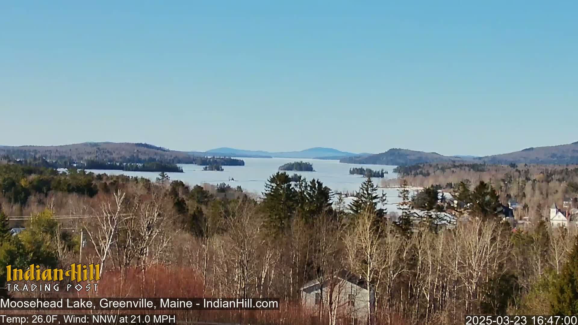 A partially frozen lake stretches to the horizon under a clear blue sky, surrounded by leafless trees and distant hills.