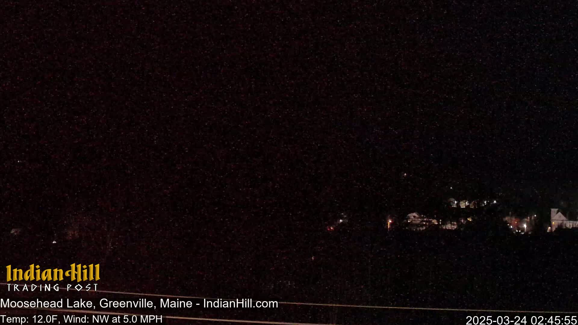 A nighttime, long shot of a dark landscape with a few scattered lights in the distance under a starlit sky, with a northwest wind at 5.0 mph and a temperature of 12 degrees Fahrenheit.