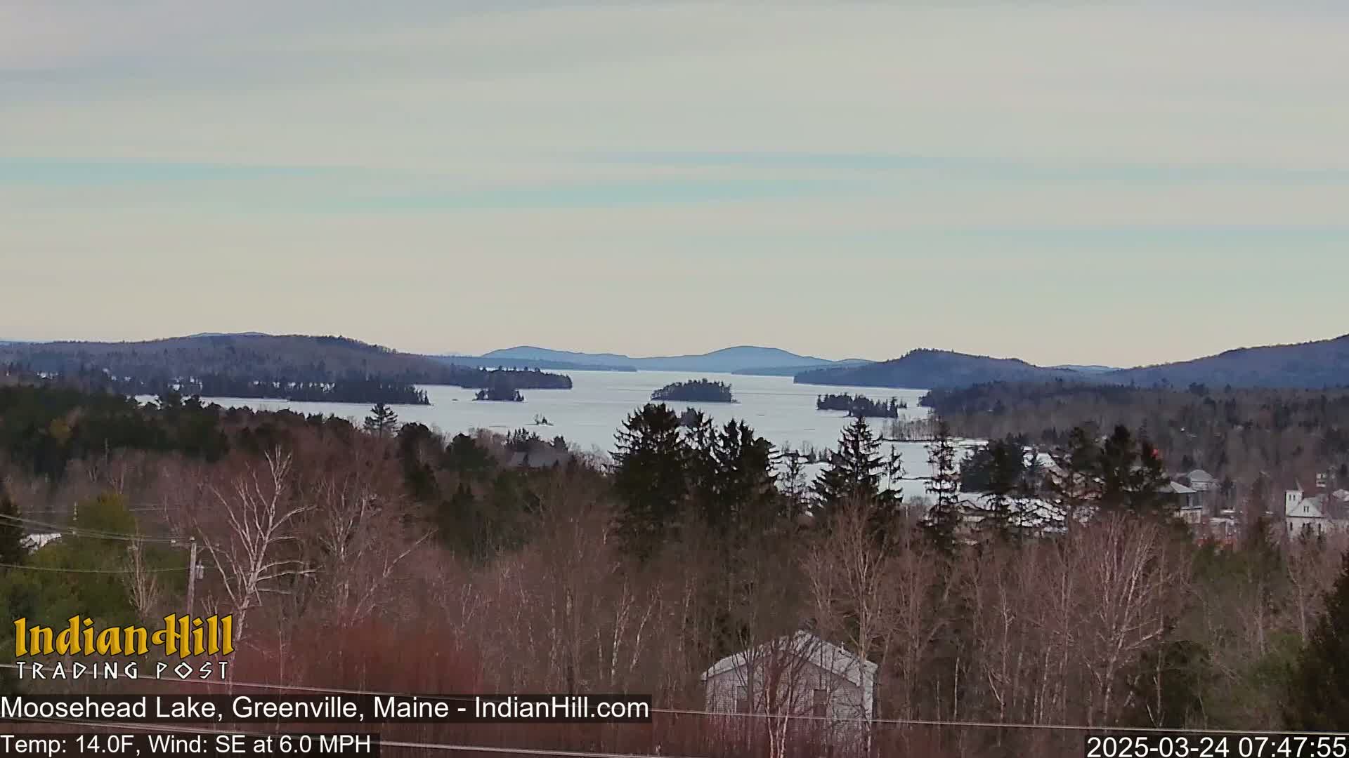 A partially frozen lake is surrounded by snow-covered trees and distant mountains under a light gray sky.