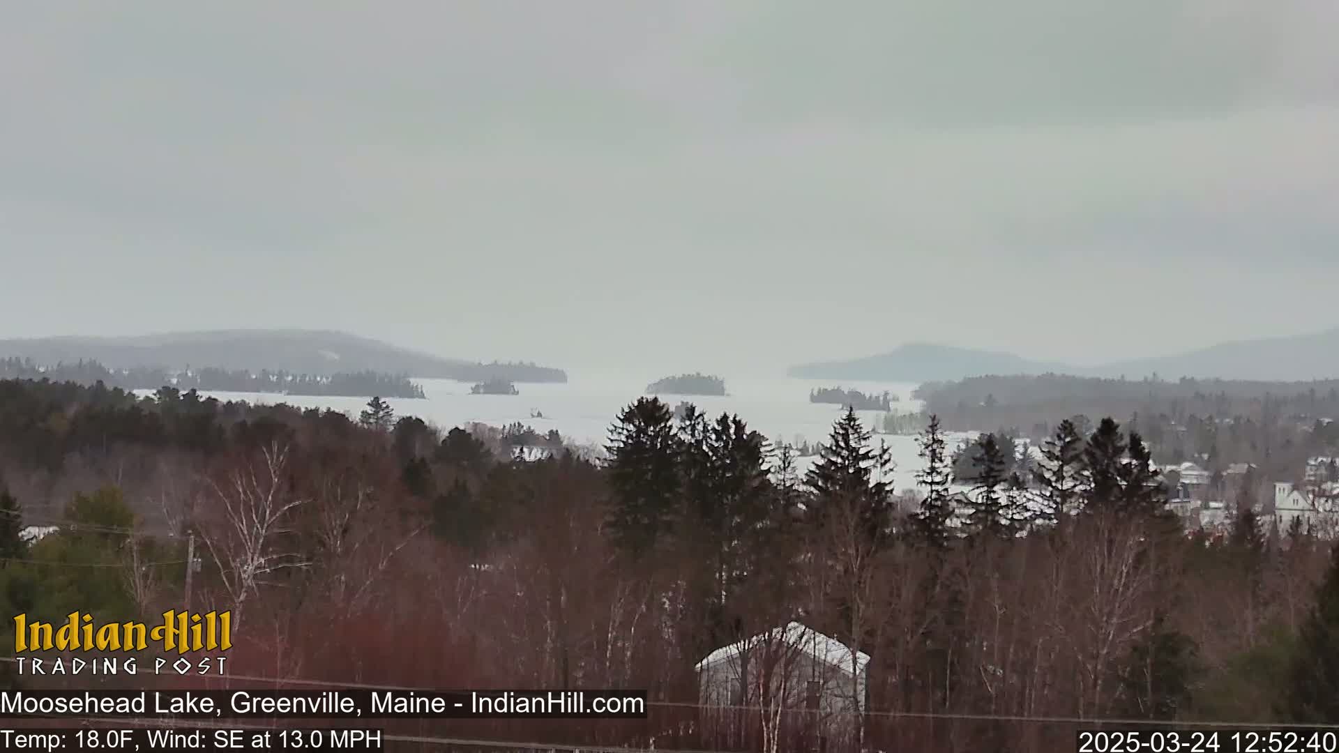 A snow-covered landscape shows a frozen lake with several islands, surrounded by a forest of dark trees under an overcast sky.