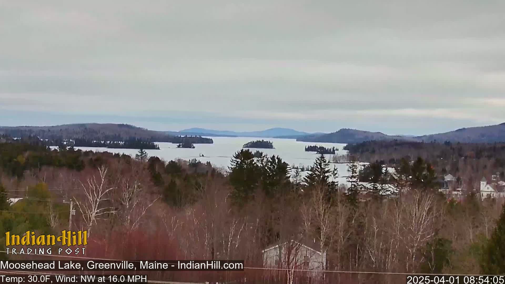 A partially frozen lake is surrounded by snow-covered hills and trees under an overcast sky.