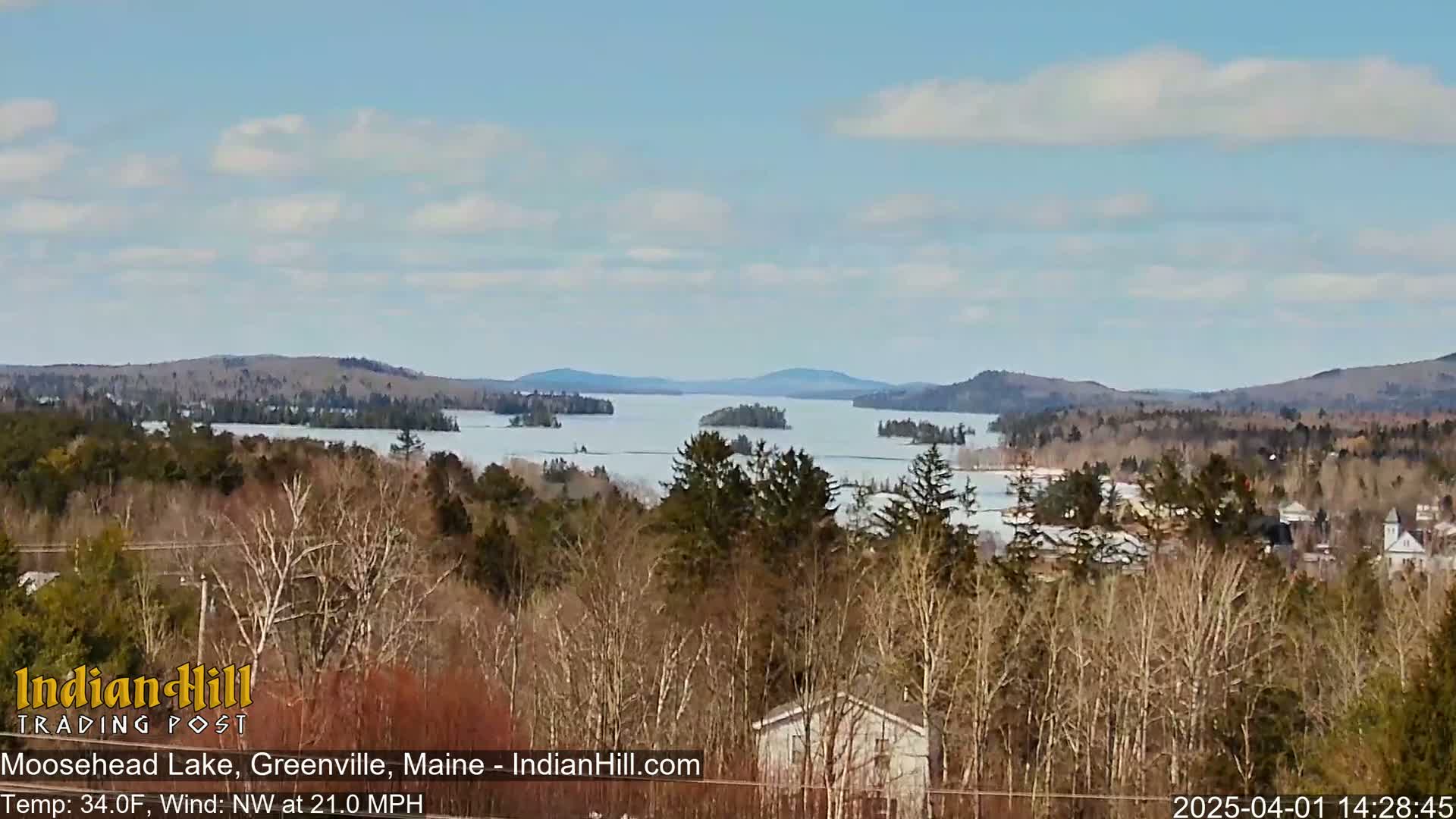 A partially frozen lake stretches into the distance under a mostly clear sky, surrounded by snow-dusted, bare trees and distant hills.