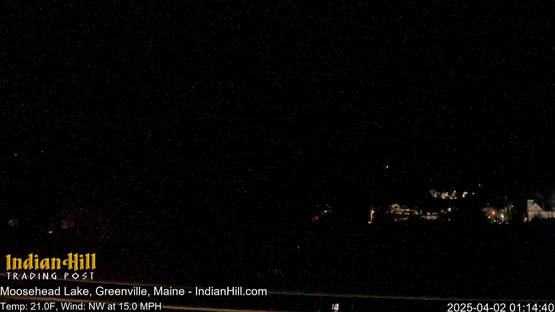 A nighttime long shot shows a dark landscape with a few distant lights under a clear, starry sky, with a northwest wind at 15 mph and a temperature of 21 degrees Fahrenheit.