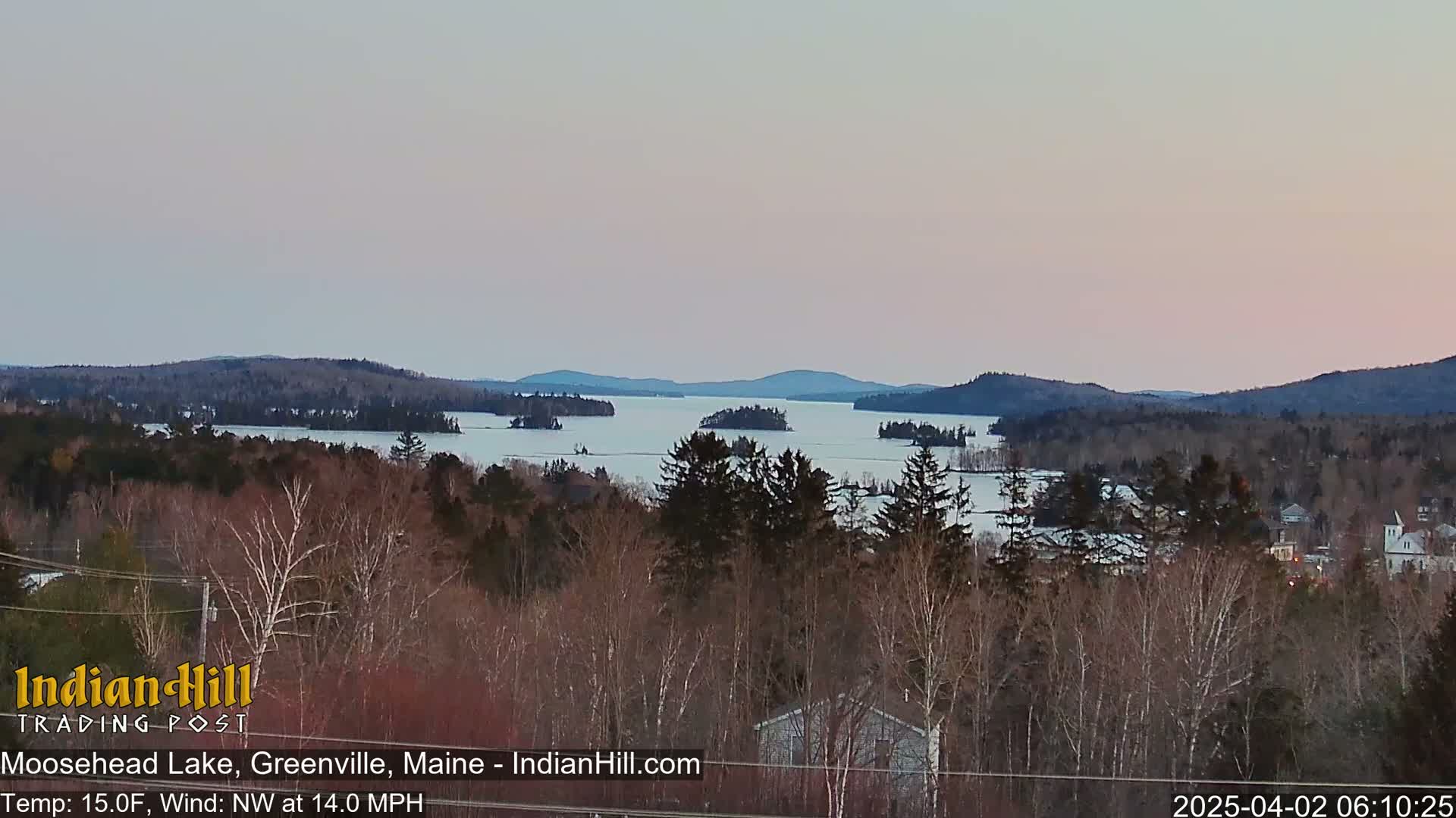 A partially frozen lake stretches to the horizon, surrounded by snow-dusted hills under a pale pink and gray sky, with a light northwest wind.