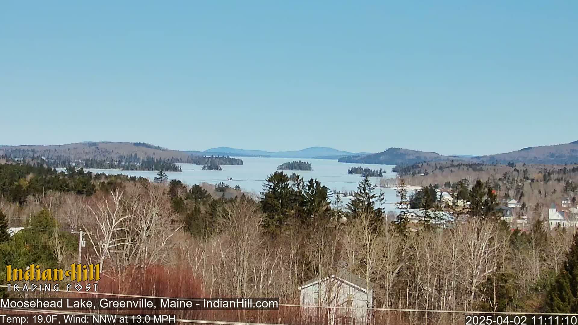 A partly frozen lake stretches to distant hills under a clear blue sky, viewed from a wooded hillside.