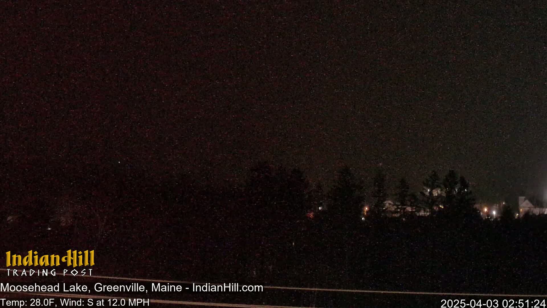 A nighttime view of dark trees under a mostly clear, starry sky with a south wind at 12 mph and a temperature of 28 degrees Fahrenheit.