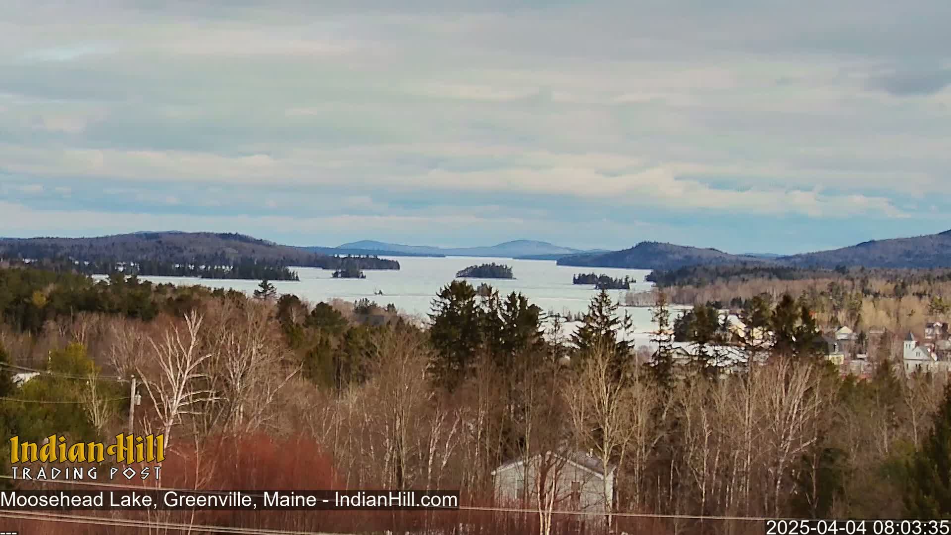 A partially frozen lake with numerous islands is seen from a distance, under a mostly cloudy sky, with a town and bare trees in the foreground.