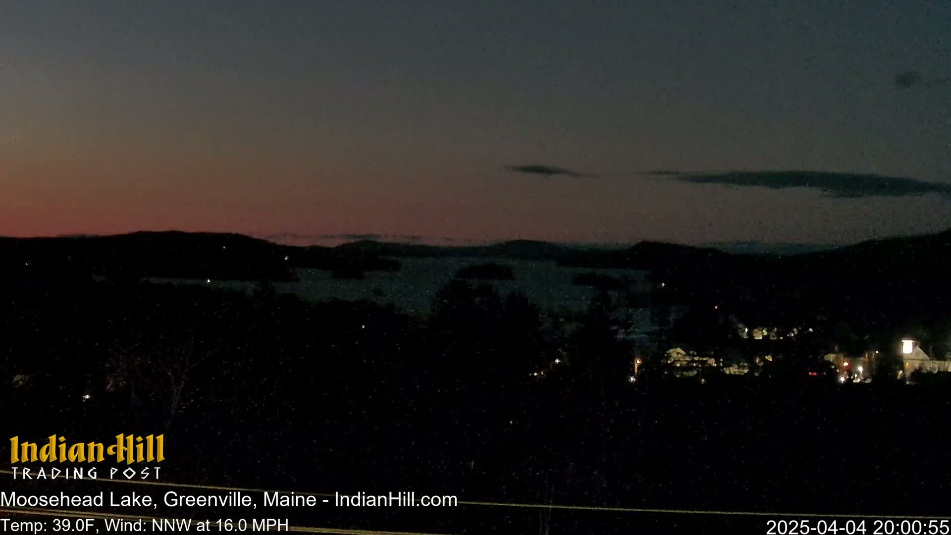 A dark, nighttime view of Moosehead Lake shows a pinkish twilight sky with a few clouds and scattered lights from a town in the foreground, under a northwest wind at 16 mph.
