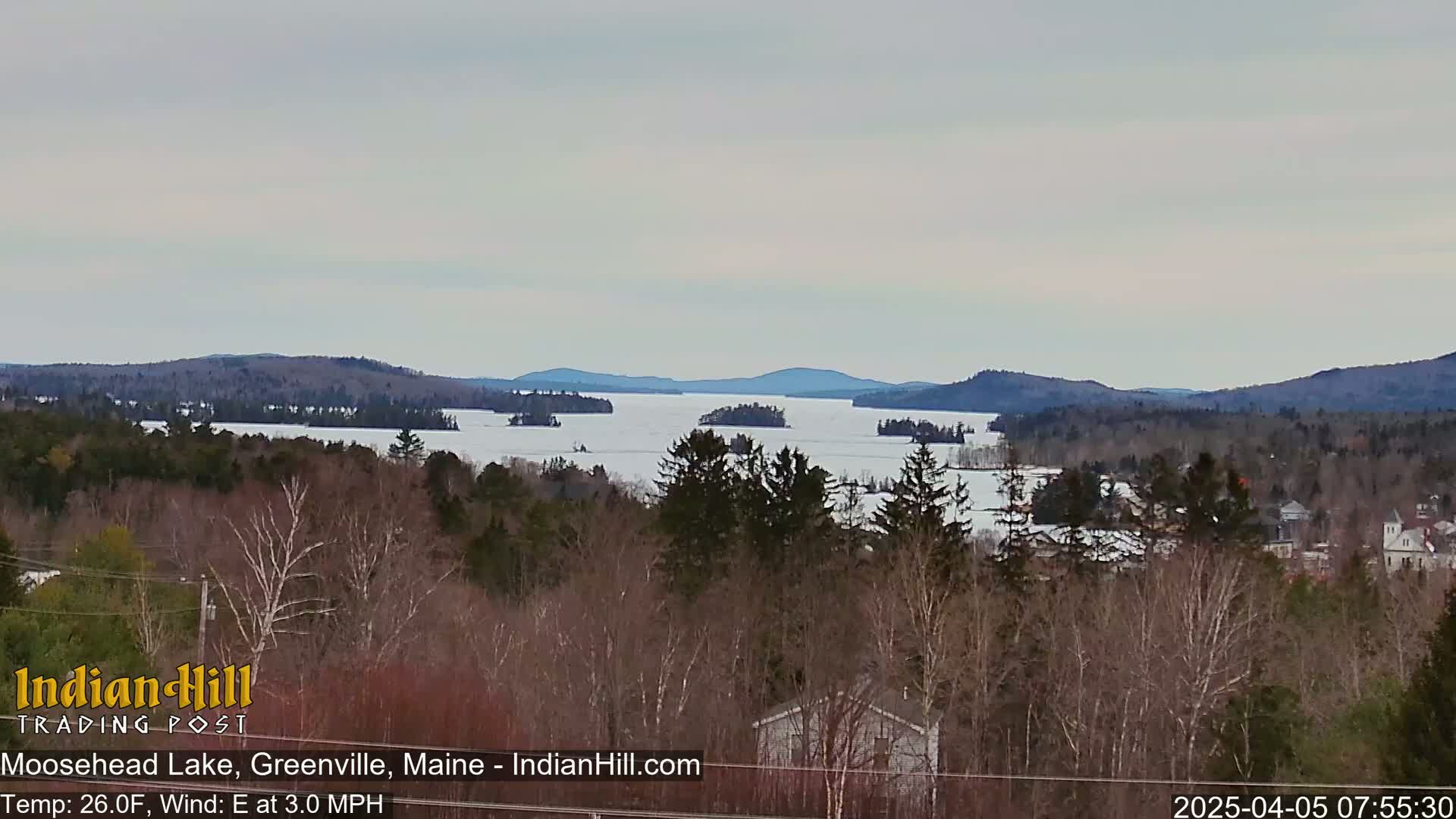 A partly frozen lake with numerous islands is visible beyond a wooded hillside under a cloudy sky.