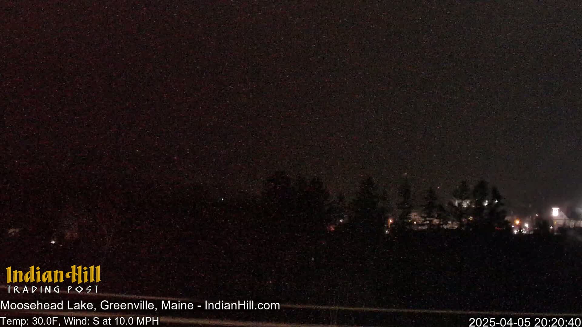 A nighttime outdoor scene showing a dark landscape with sparsely lit trees and buildings under snowy conditions.