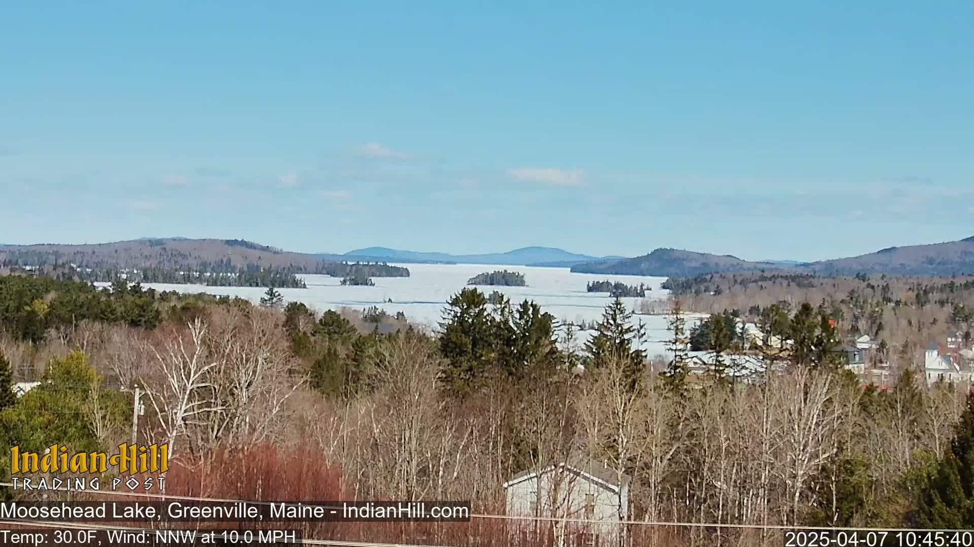 A partially frozen lake stretches to distant mountains under a clear blue sky, with bare trees in the foreground.