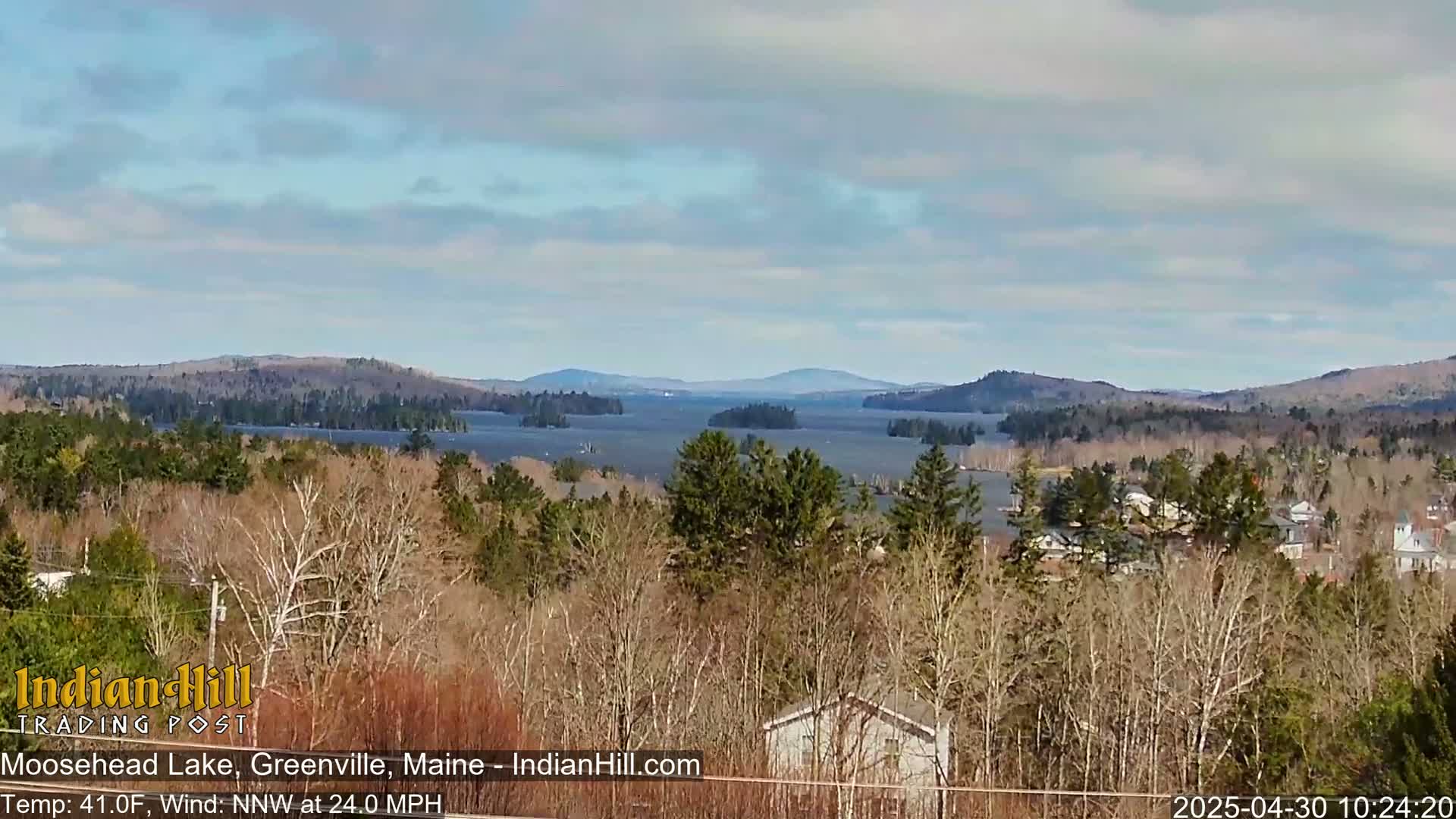 A partly cloudy day reveals a view of a large lake surrounded by hills and sparsely wooded areas, with some houses visible in the foreground.